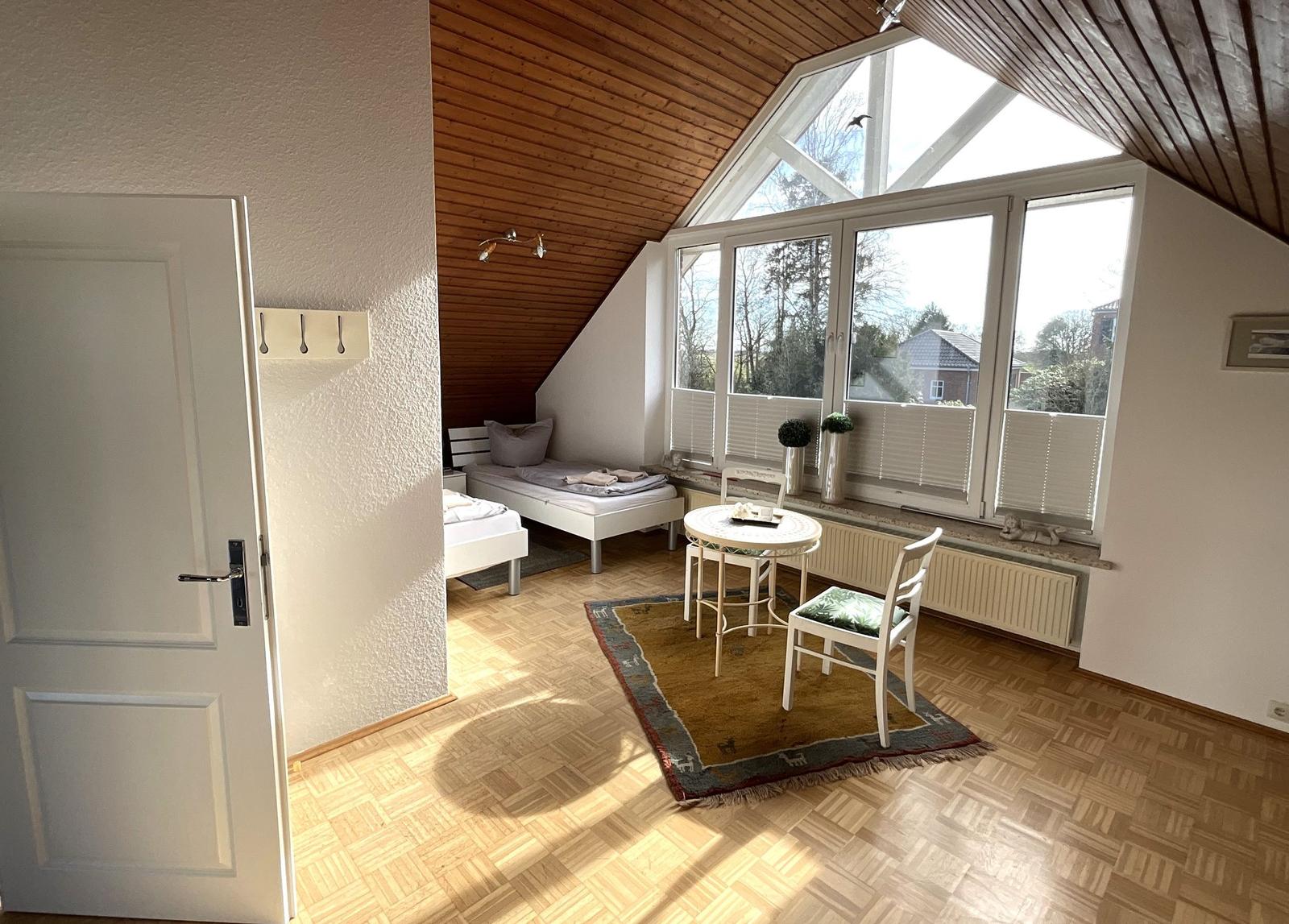 Zimmer mit Dachschrägen, Fenster und Holzboden. Tisch, Stühle und Bett sind vorhanden.