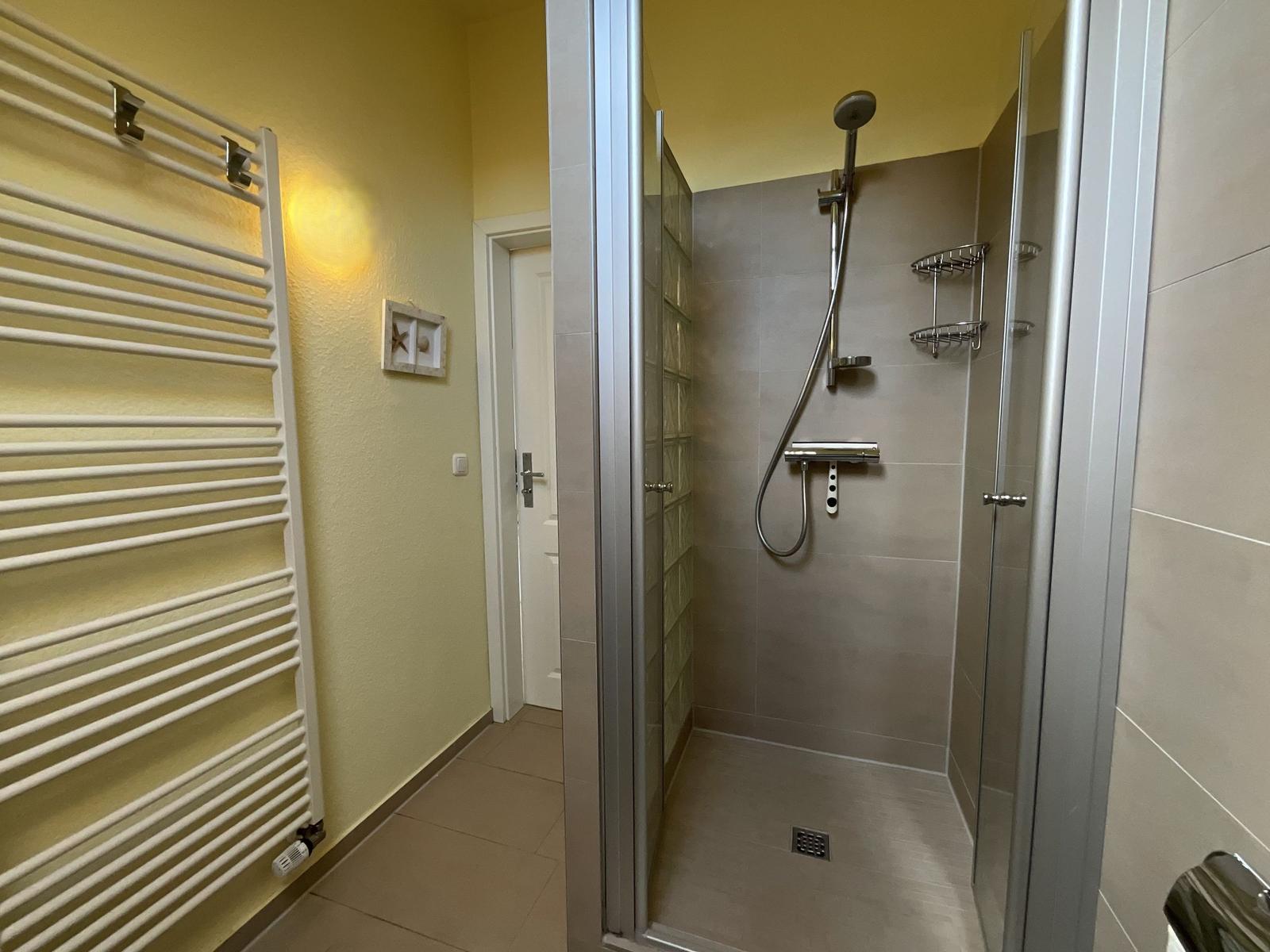 Dusche mit Glaswand, Duschbrause und Ablage, daneben Heizkörper an gelber Wand.