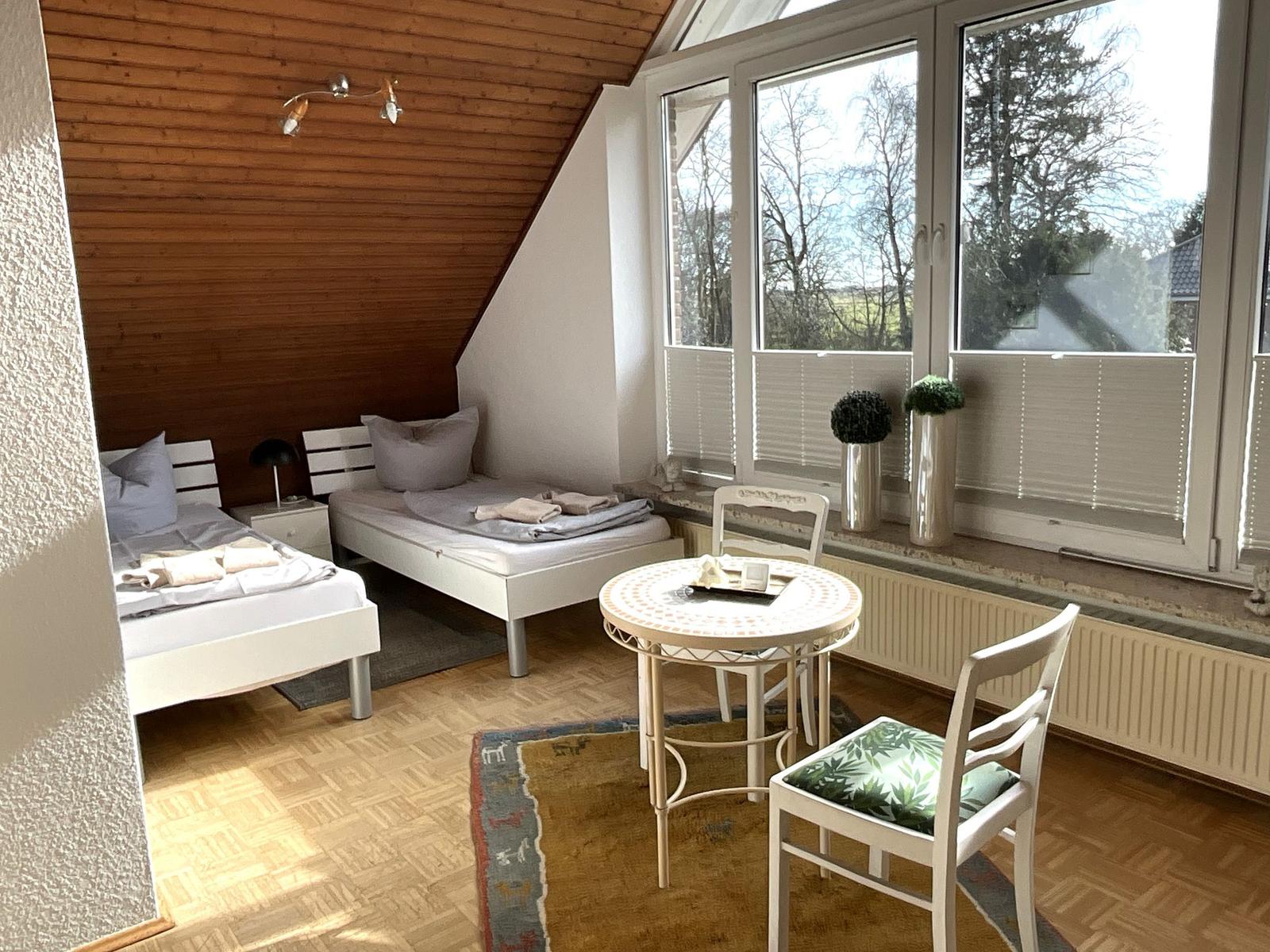 Zimmer mit zwei Betten, Tisch und Stühlen, großem Fenster und Holzdecke.