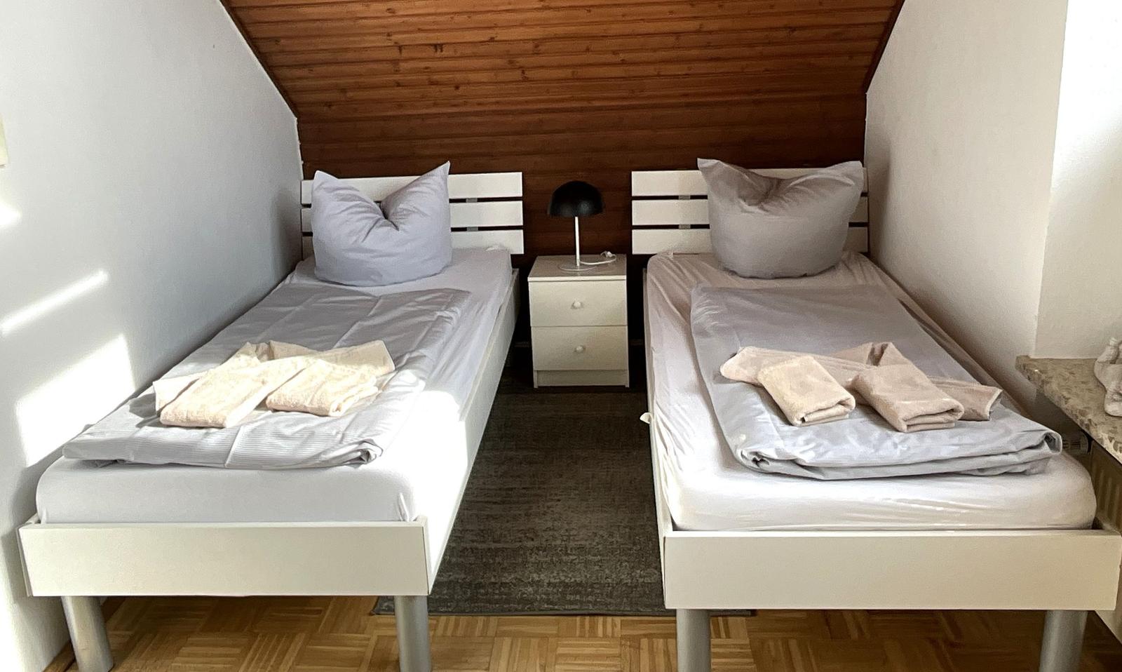 Zwei Einzelbetten mit Kissen und Handtüchern. Zwischen ihnen steht ein Nachttisch mit Lampe.