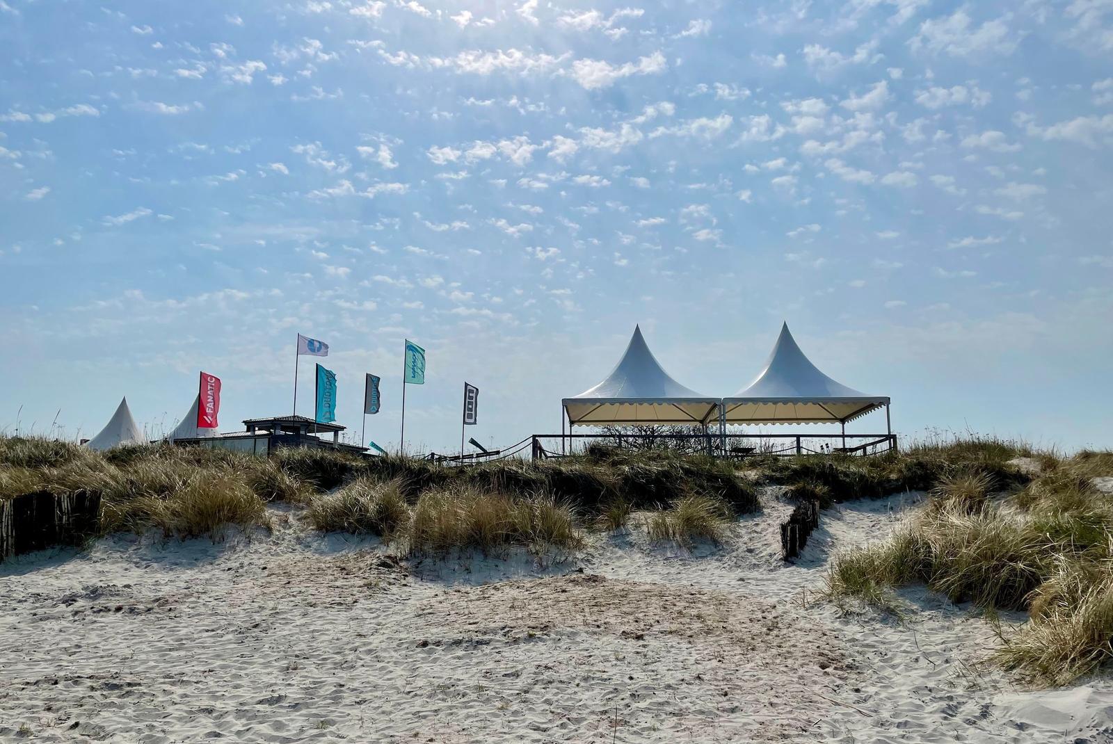 Strand mit Dünen, weißen Zelten und Flaggen unter blauem Himmel.