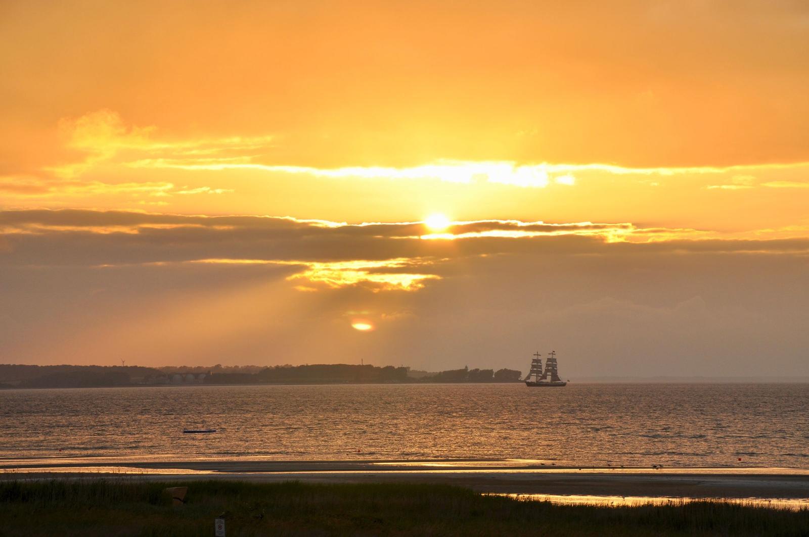 Sonnenuntergang über dem Meer mit Segelschiff und Strand im Vordergrund.