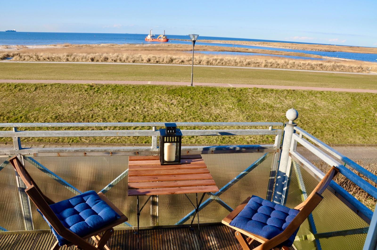 Terrasse mit Tisch und zwei Stühlen mit blauen Kissen. Blick auf Grünfläche und Meer.