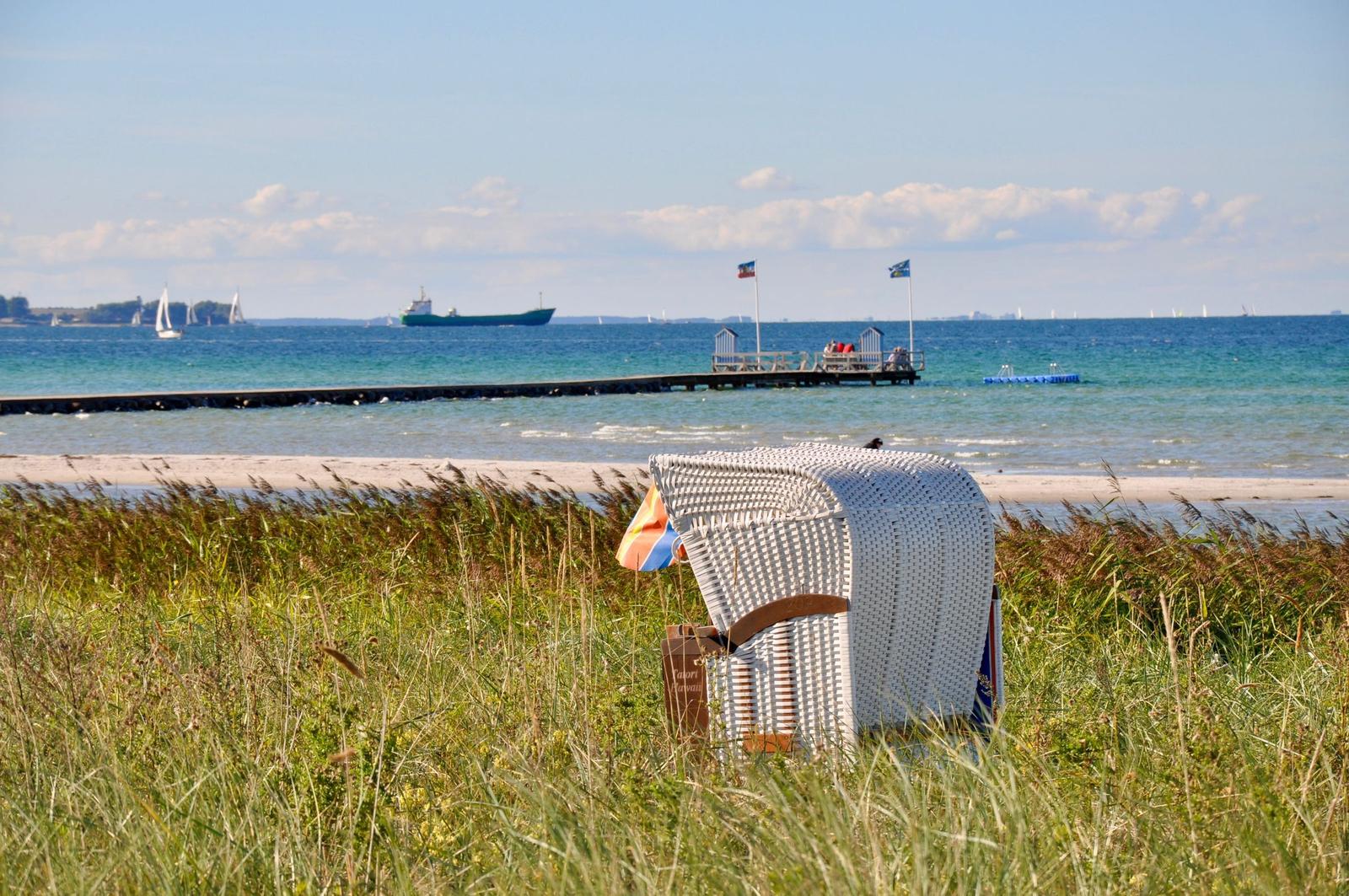 Weißer Strandkorb steht im Gras. Im Hintergrund See, Pier und Schiffe.