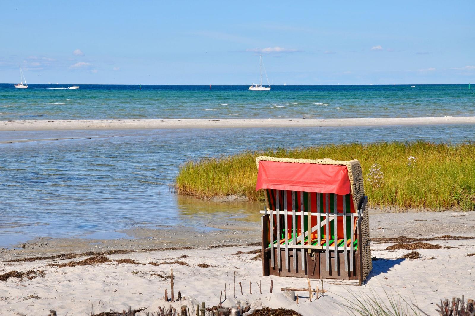 Ein roter Strandkorb steht am sandigen Ufer. Im Hintergrund sind Segelboote auf dem Meer zu sehen.