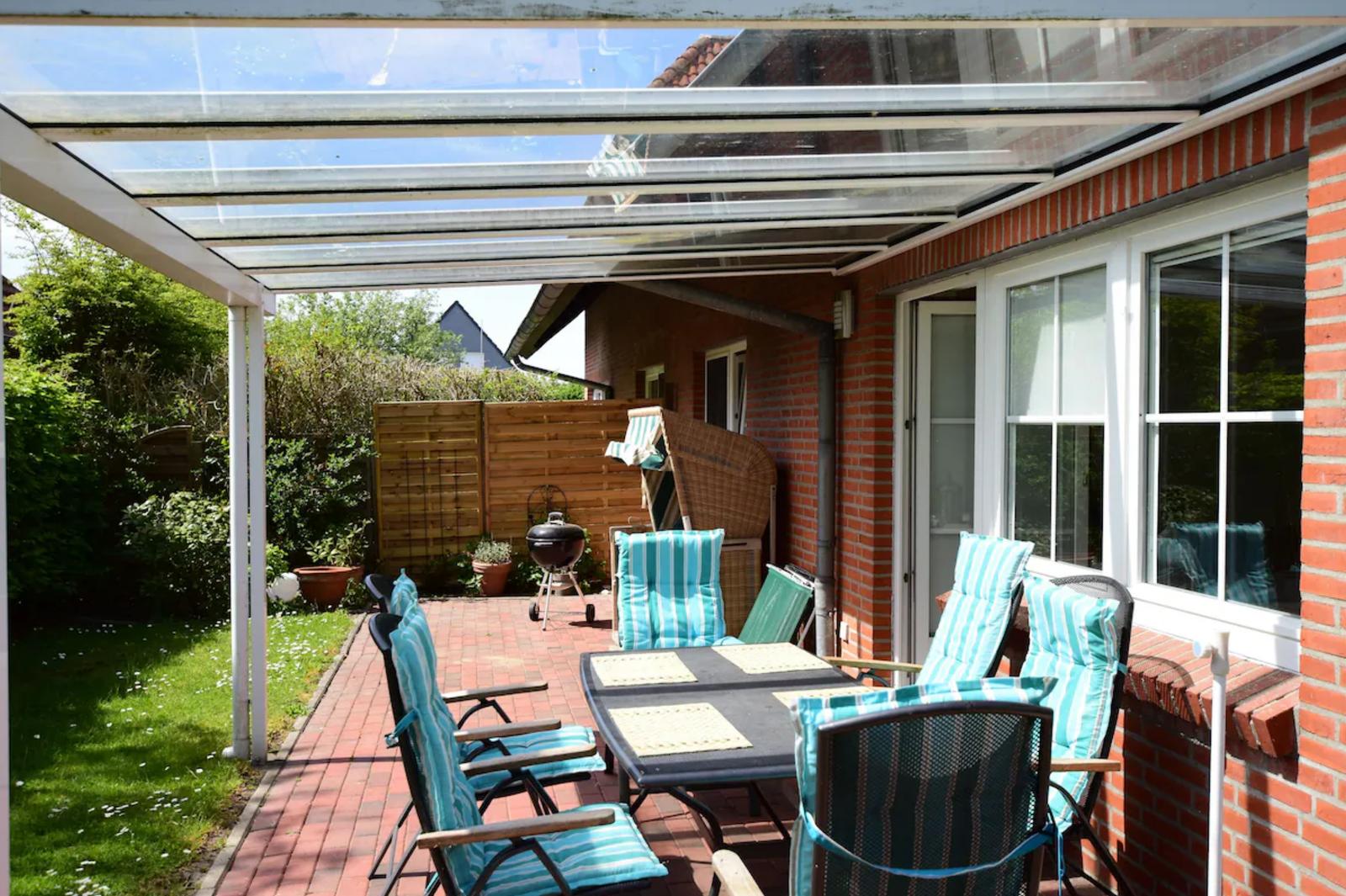 Terrasse mit Sitzgruppe, Grill und Glasdach. Roter Kiesweg und grüner Rasen.