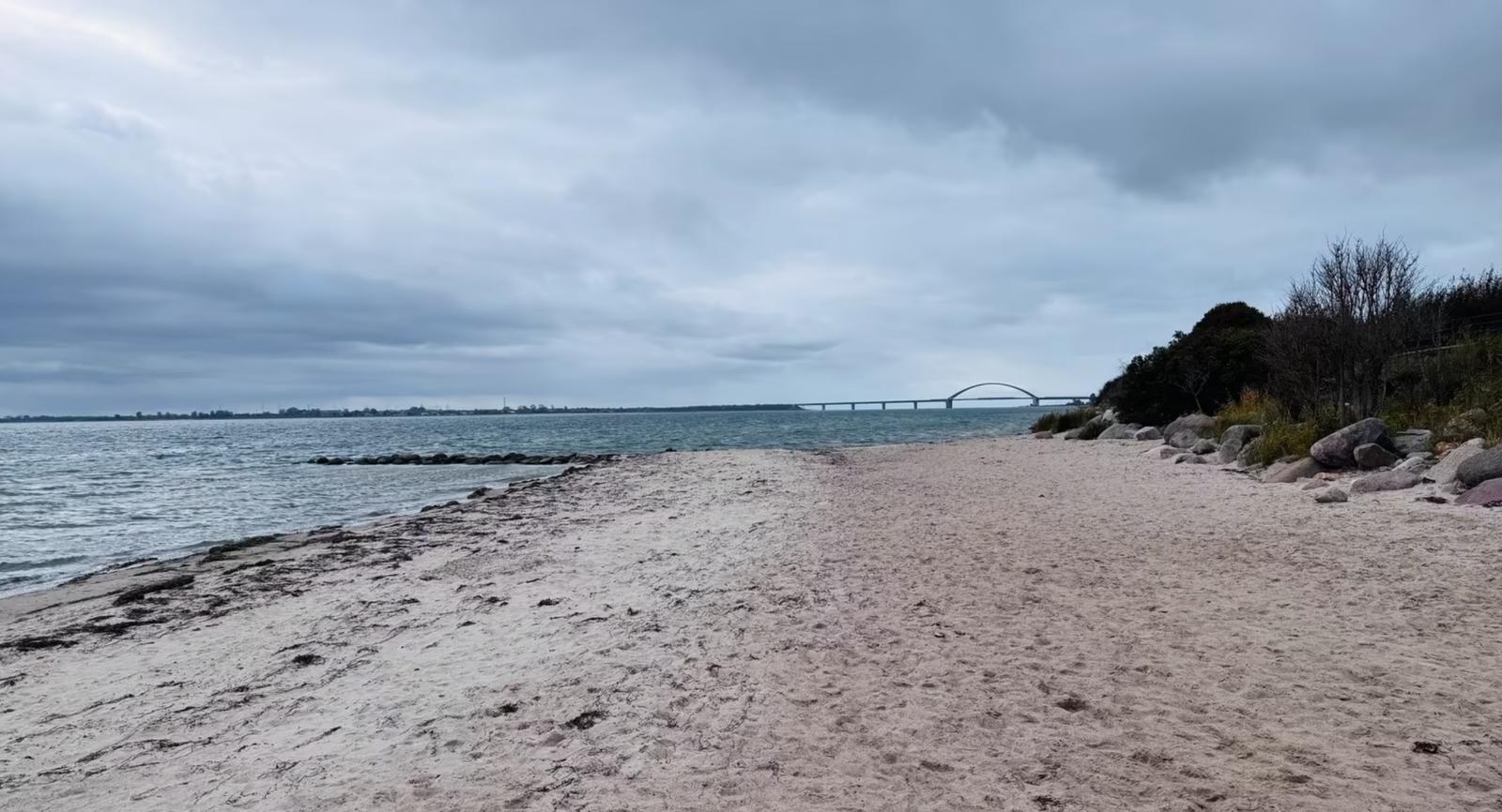 Strand mit Sand, Felsen und Brücke im Hintergrund unter bewölktem Himmel