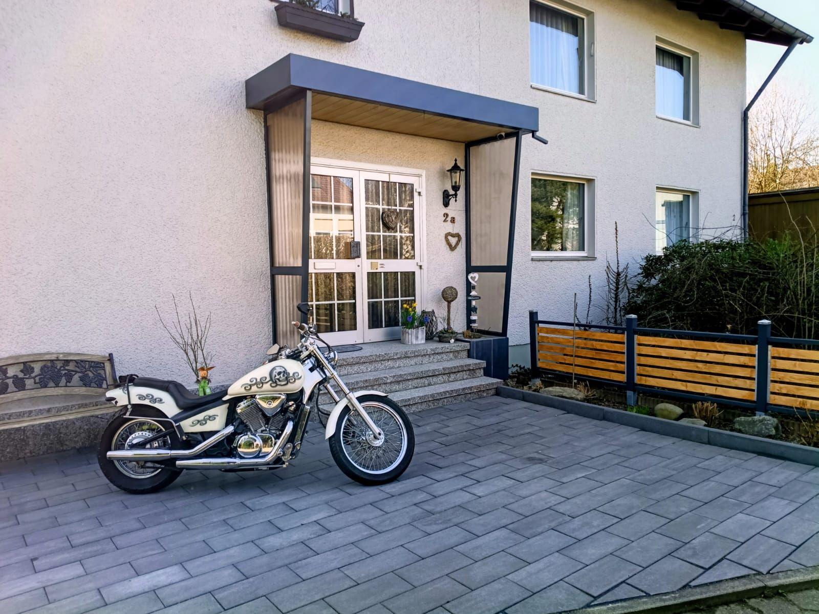 Eine weiße Motorrad steht vor der Eingangstür eines Hauses mit gepflastertem Hof.