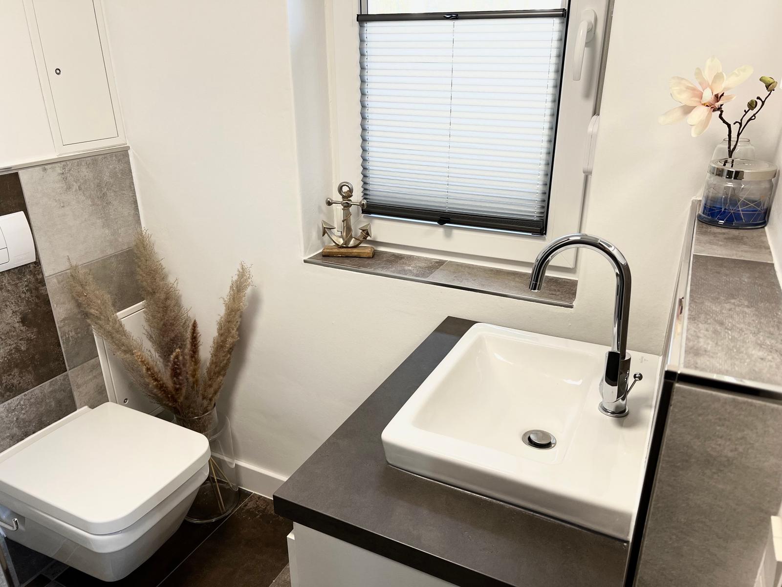 Badezimmer mit Waschbecken, Toilette und Fenster mit Jalousie.