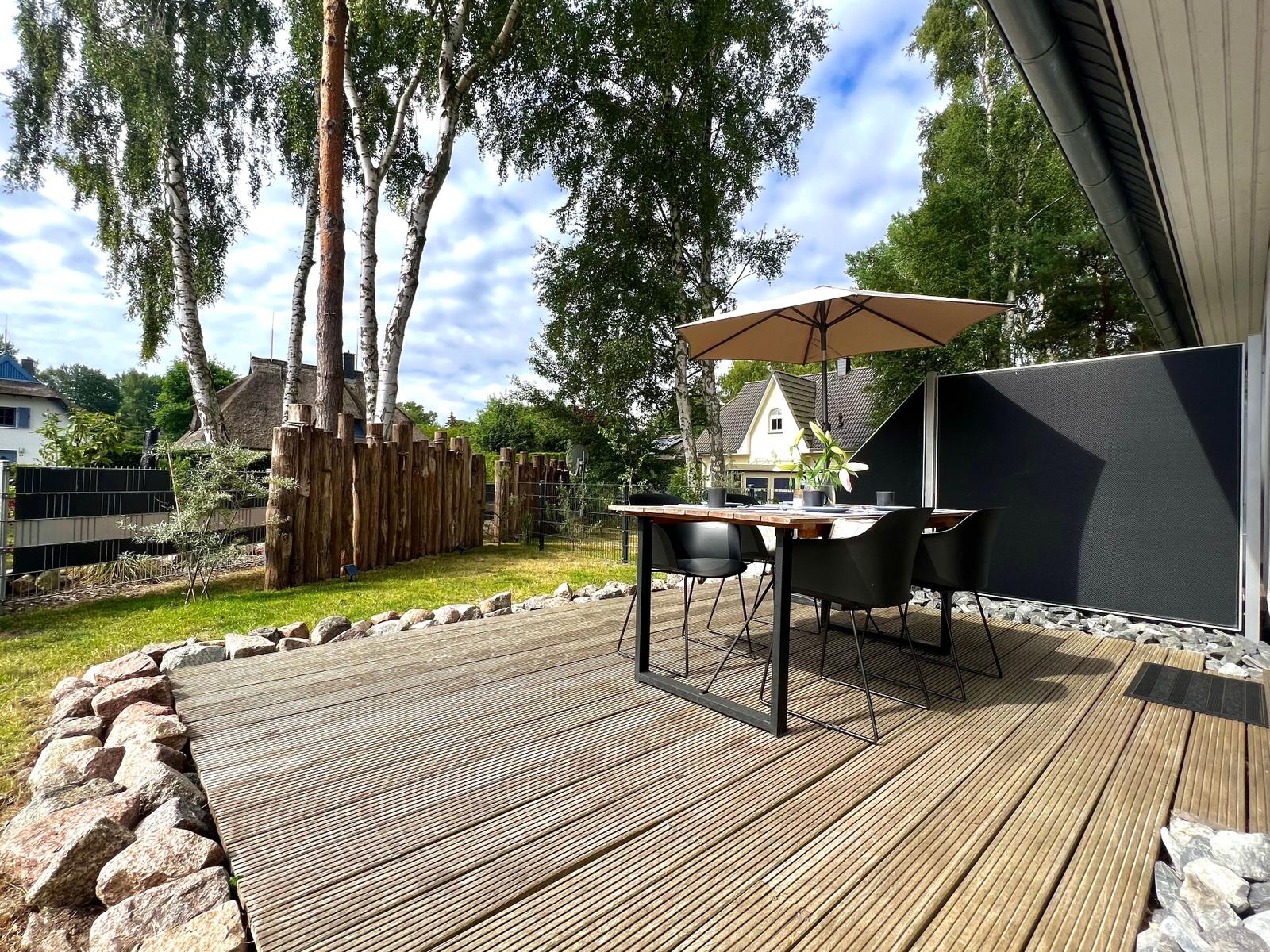 Holzterrasse mit Tisch, Stühlen und Sonnenschutz unter Bäumen.