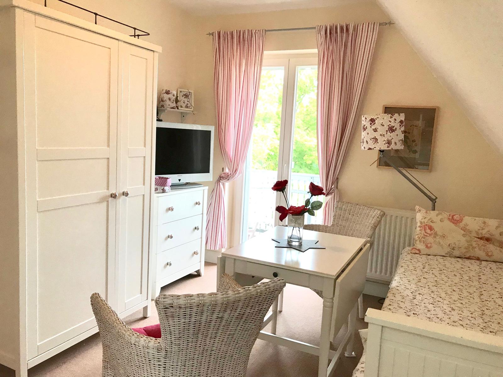 Schlafzimmer mit Bett, Tisch, Stühlen und TV. Fenster mit rosa Vorhängen.