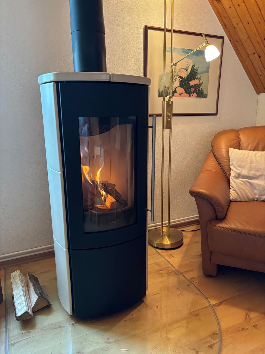 Kaminofen mit brennendem Holz, braunes Ledersofa und Stehlampe in einem Raum mit Holzboden und schräger Decke.