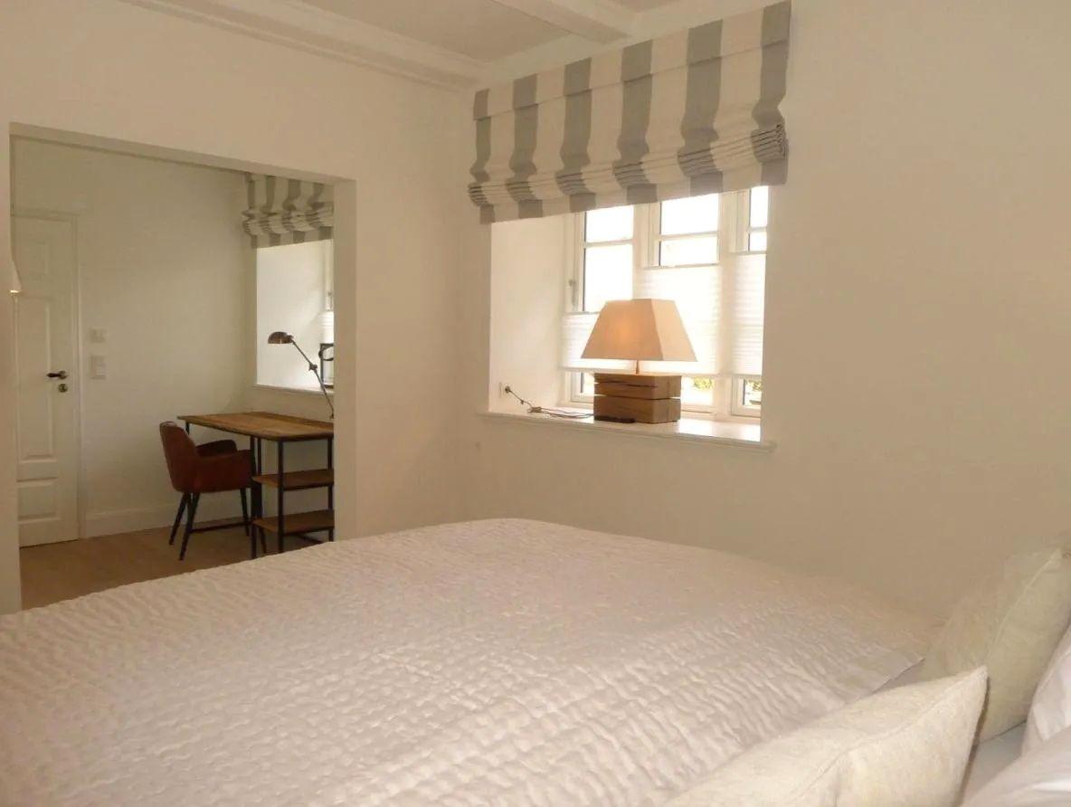 Schlafzimmer mit Bett, Fenster und Schreibtisch im Hintergrund.