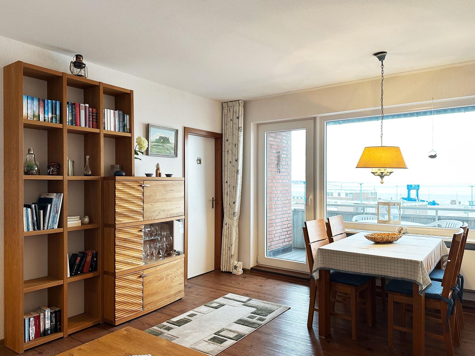 Esszimmer mit Tisch, Stühlen und Blick auf Balkon. Holzregal mit Büchern und Glasvitrine.