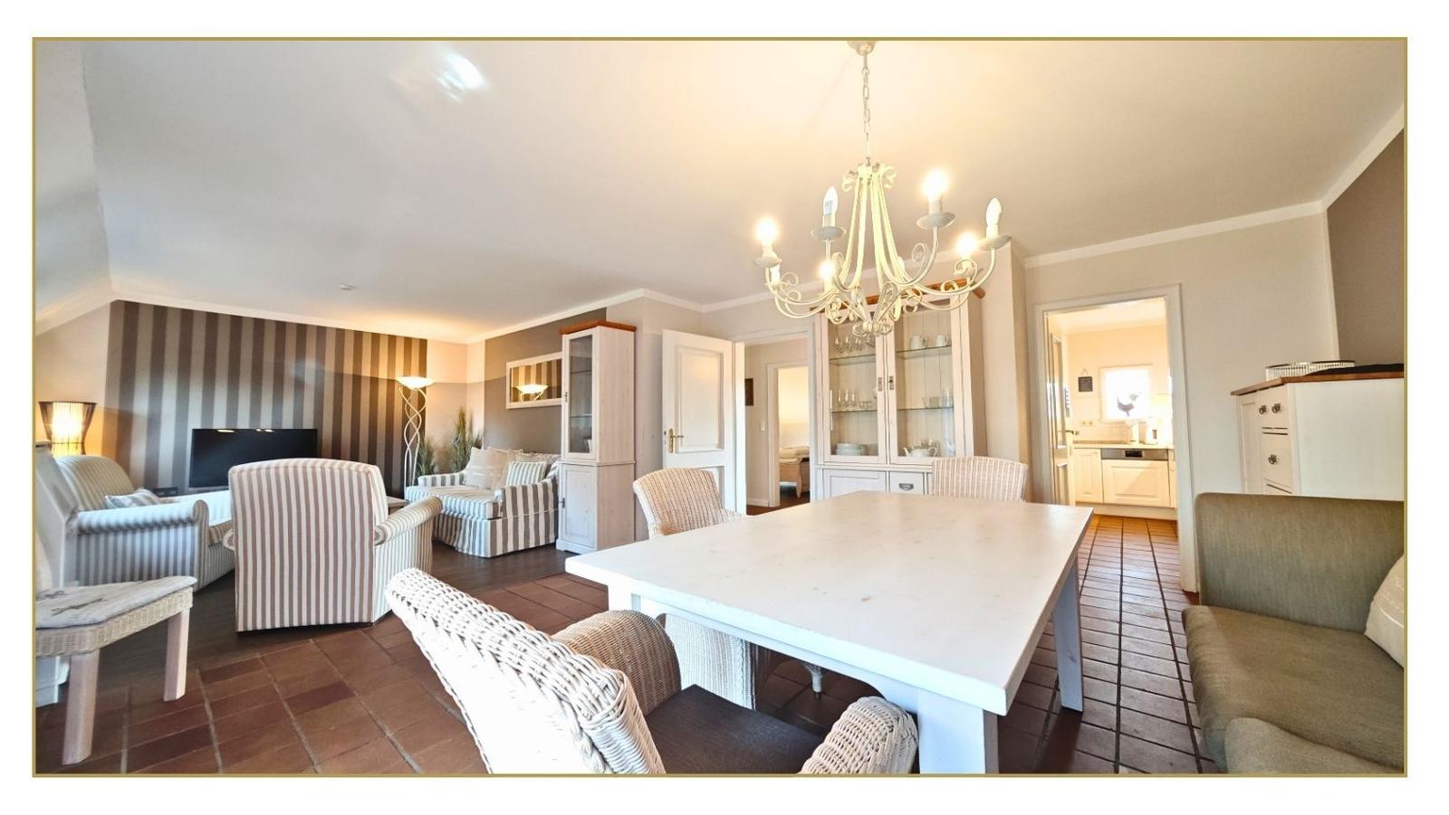 Großes Esszimmer mit weißem Tisch und Stühlen. Wohnbereich mit Sofa und Fernseher. Küche im Hintergrund.