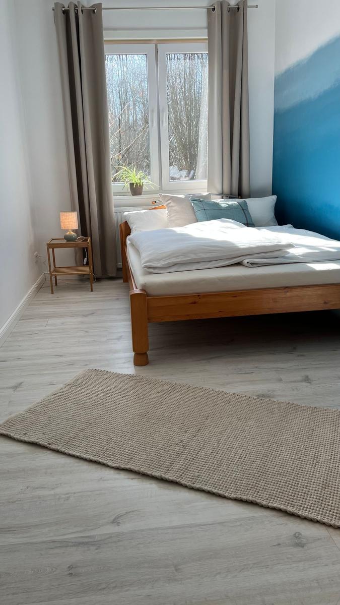 Schlafzimmer mit Holzbett, Fenster und Teppich auf hellen Bodenfliesen.