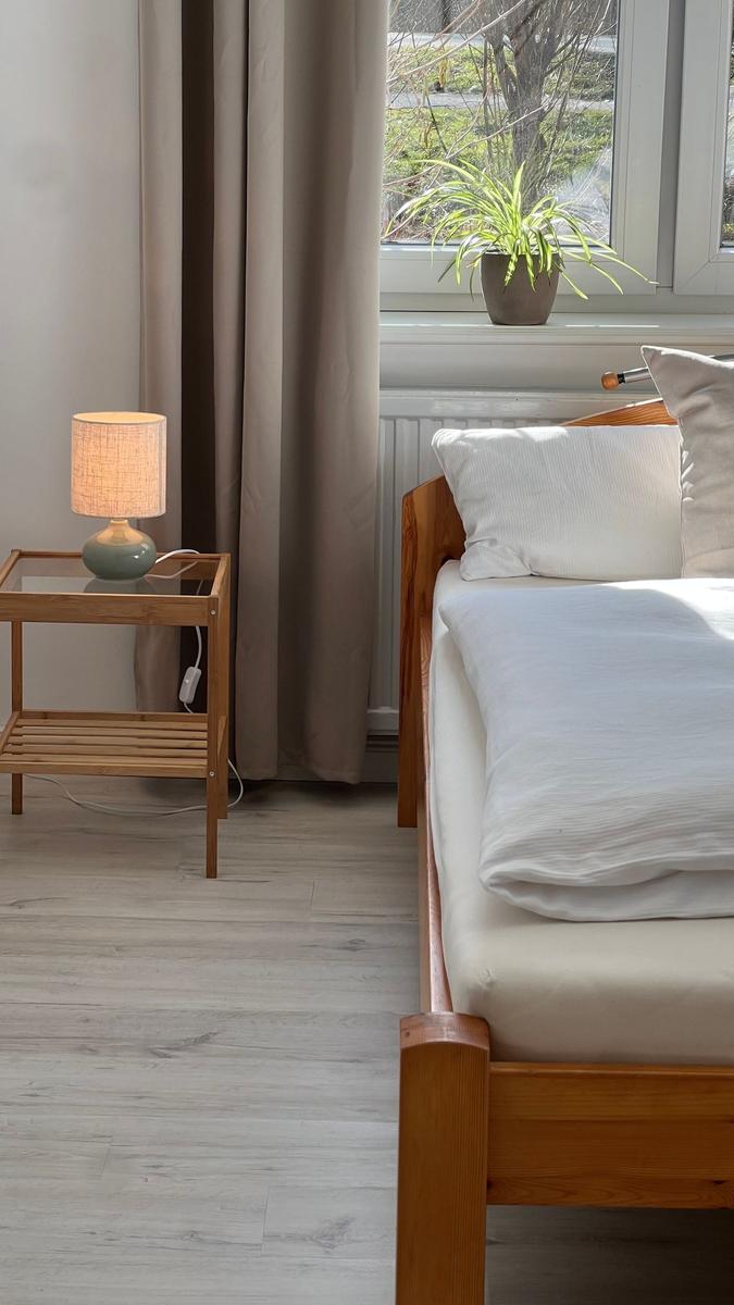 Schlafzimmer mit Bett, Nachttisch und Pflanze am Fenster.