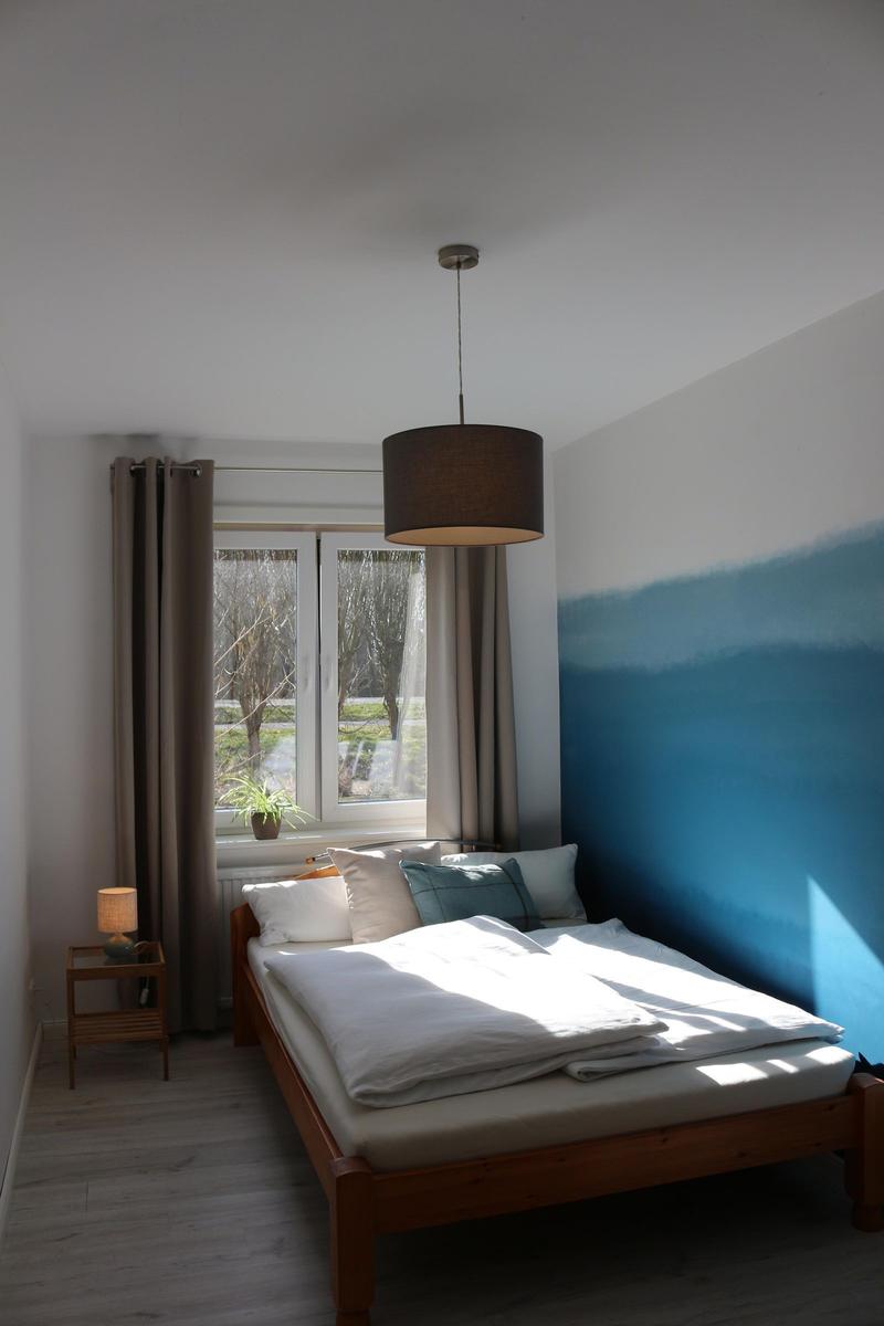Schlafzimmer mit Holzbett, Fenster und blauer Wand. Hängelampe und Nachttisch.