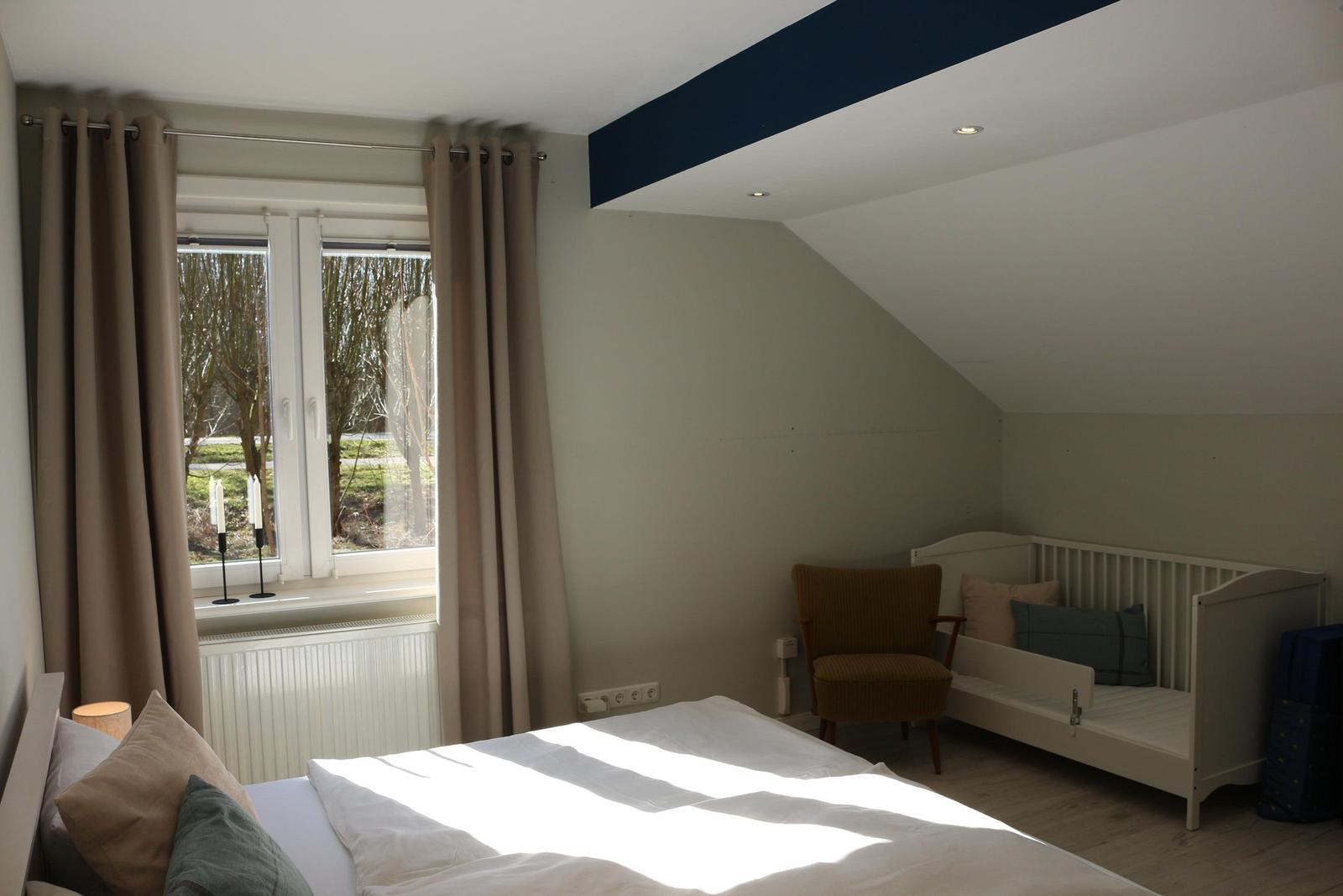 Schlafzimmer mit Bett, Kinderbett und Fenster. Dachgeschoss mit Holzboden und dunkler Deckenleiste.