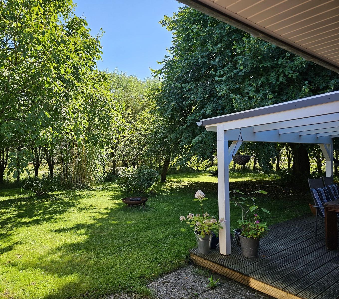 Terrasse mit Sitzgelegenheiten und Pflanzen. Hintergrund: grüner Garten mit Bäumen und Feuerkorb.