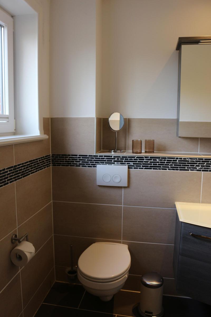 Badezimmer mit Toilette, Waschbecken und Spiegel. Wandfliesen in Braun- und Schwarzton.