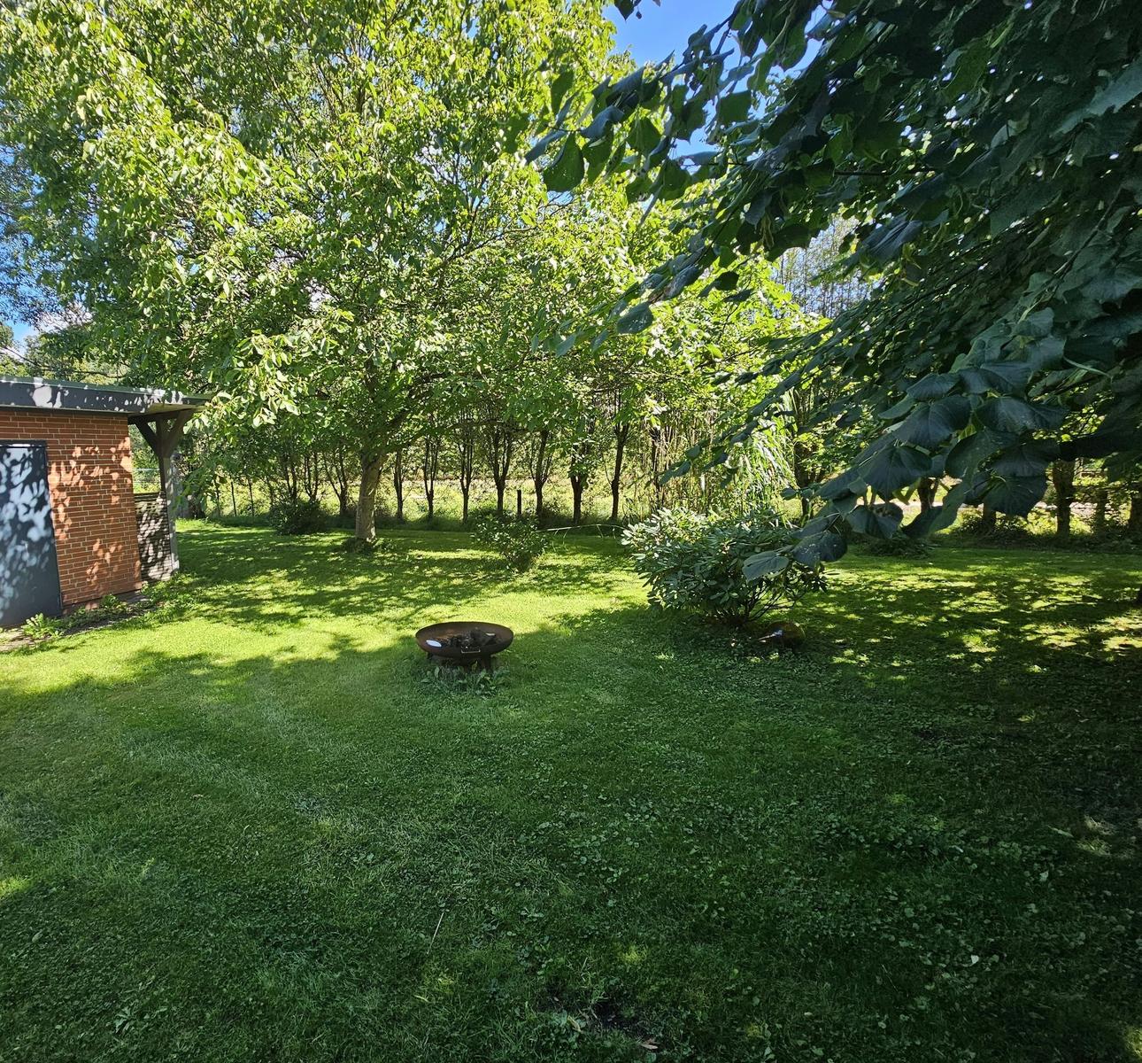 Grüner Garten mit Feuerkorb und Bäumen. Links steht ein kleines Gebäude.