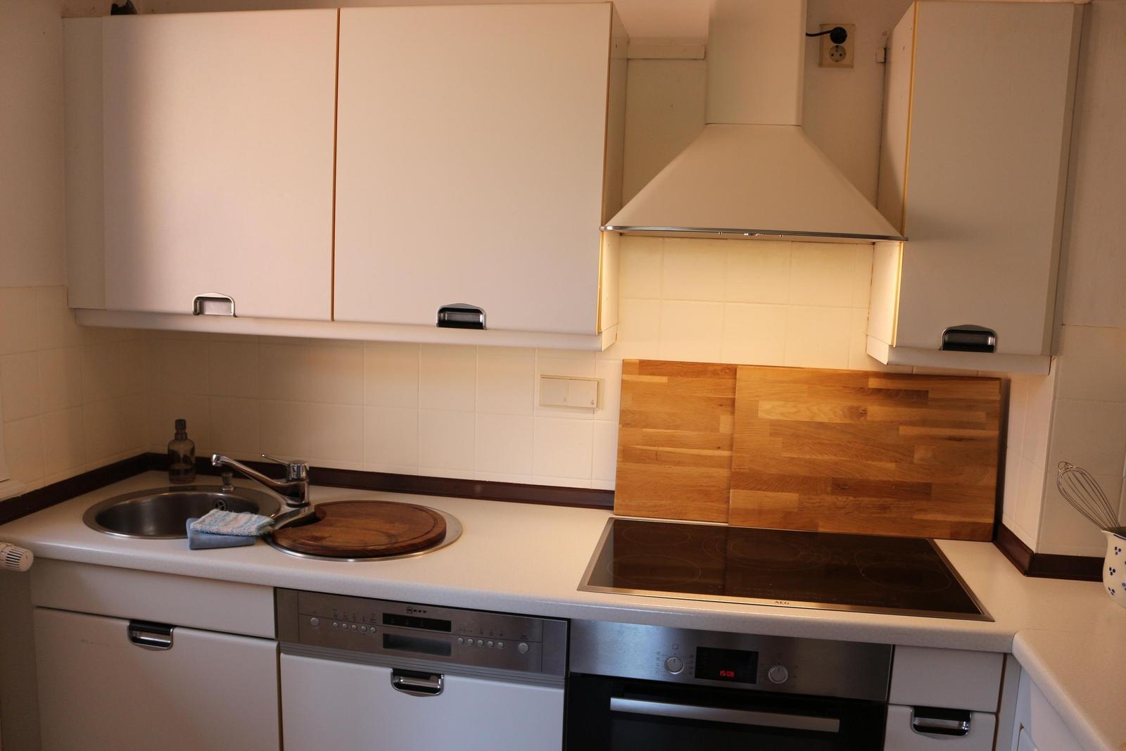 Küche mit Spüle, Kochfeld und Backofen. Holzrückwand und weiße Schränke.