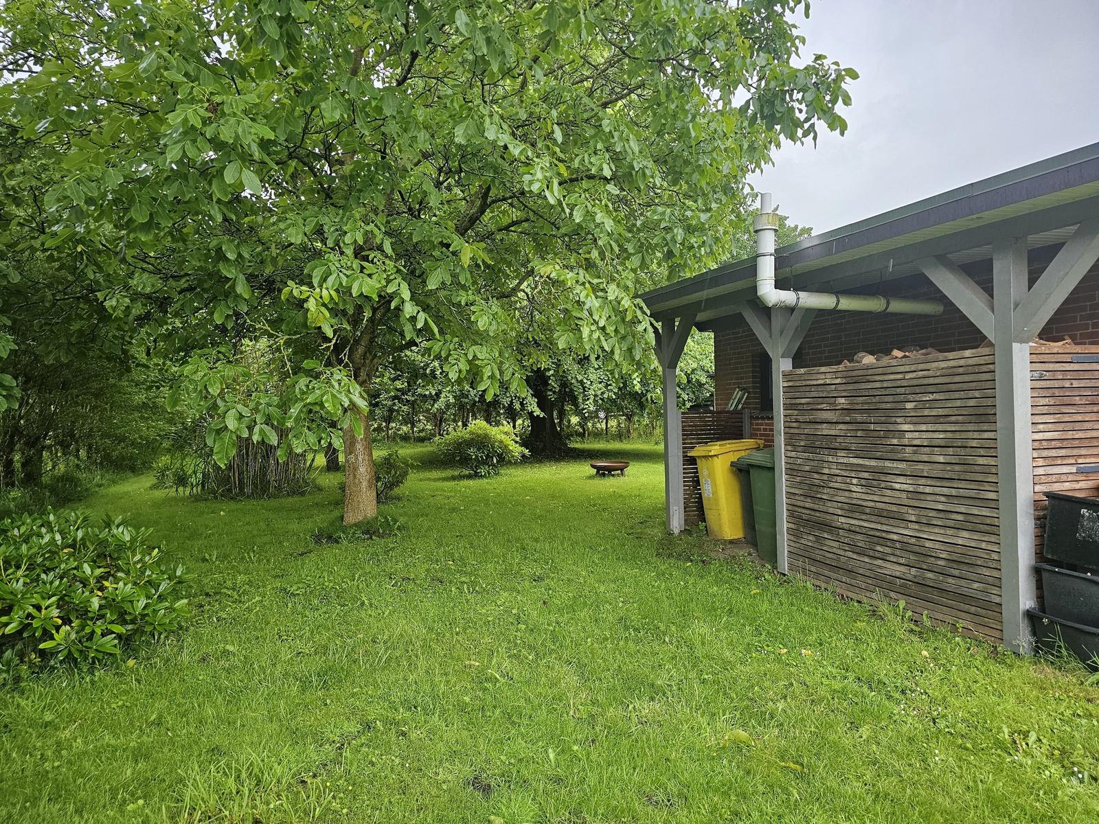 Grüner Garten mit Baum, Holzverkleidung und Müllcontainern unter Dach.
