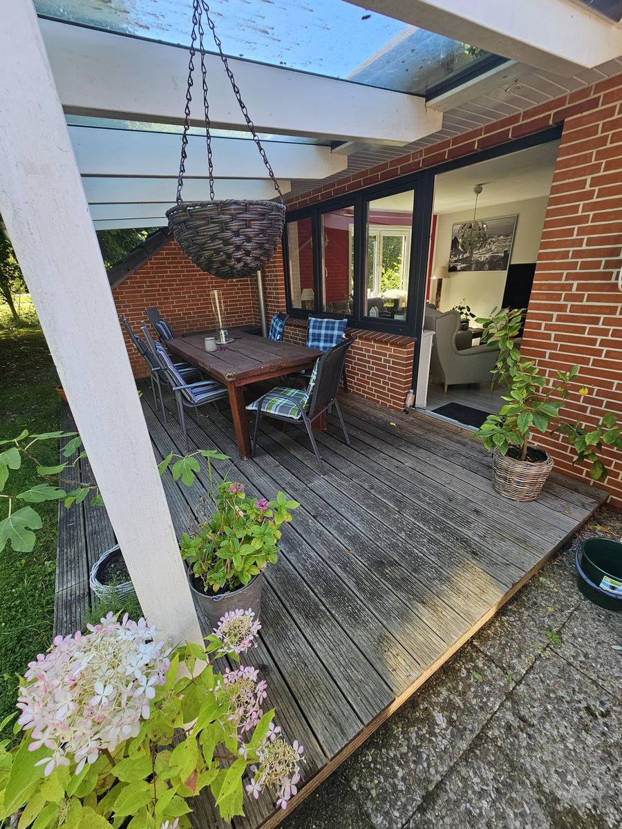 Terrasse mit Tisch, Stühlen und hängender Pflanze. Durchgang ins Haus mit Blick in Wohnzimmer.