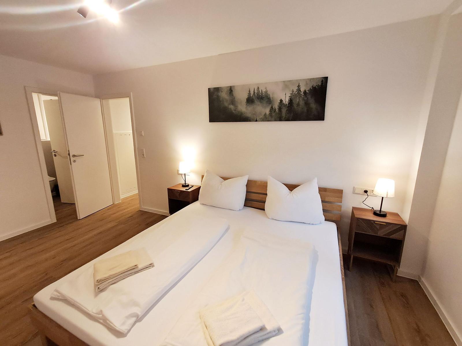 Schlafzimmer mit Doppelbett, zwei Nachttischen und Wandbild. Holzboden und angrenzender Badraum.