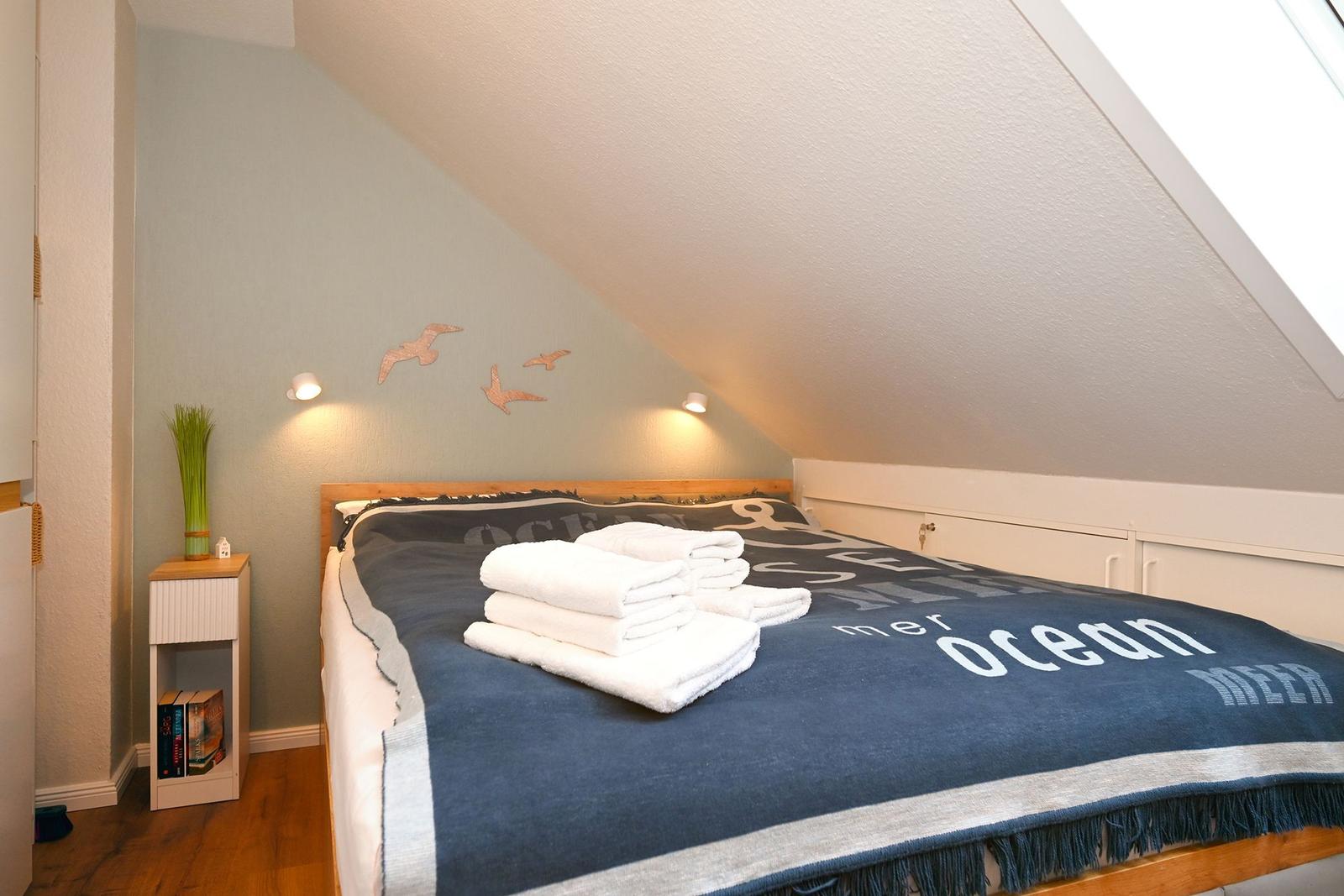 Schlafzimmer mit Dachschrägen, Bett mit Decke und Handtüchern, Nachttisch mit Pflanze und Büchern.