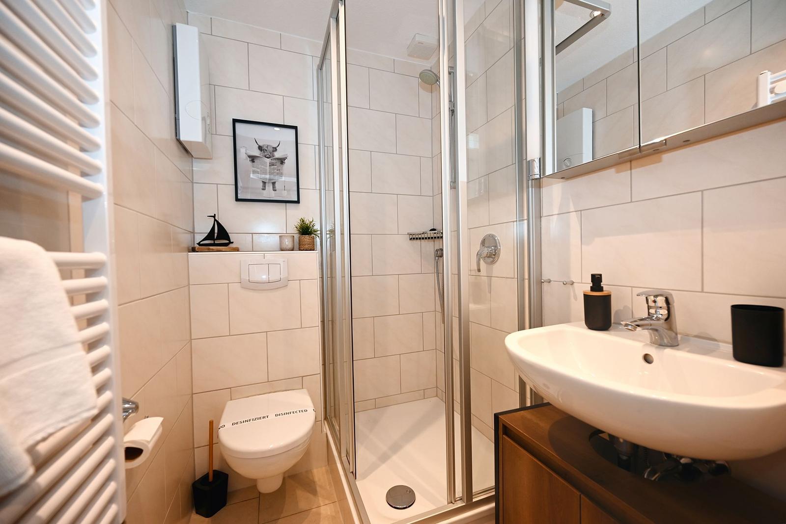 Badezimmer mit Dusche, Waschbecken und Toilette. Weiße Fliesen und Holzunterschrank.