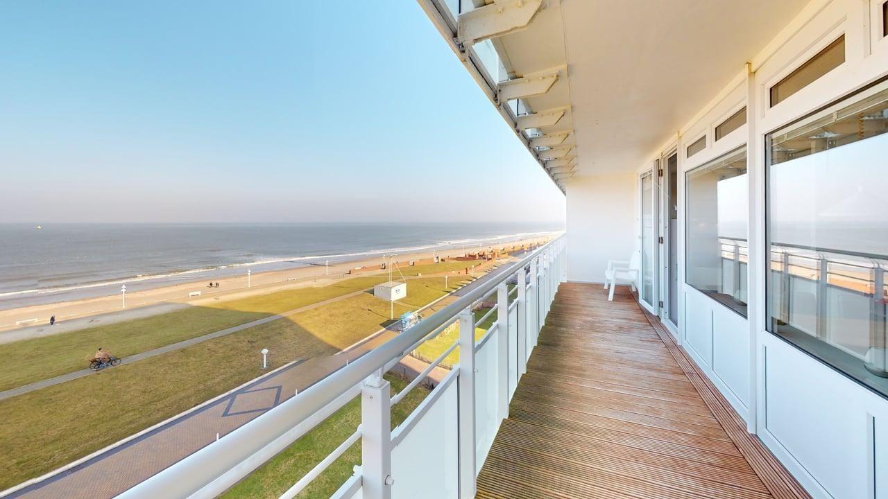 Terrasse mit Meerblick, Holzboden und weißer Brüstung. Blick auf Strand und Grünfläche.