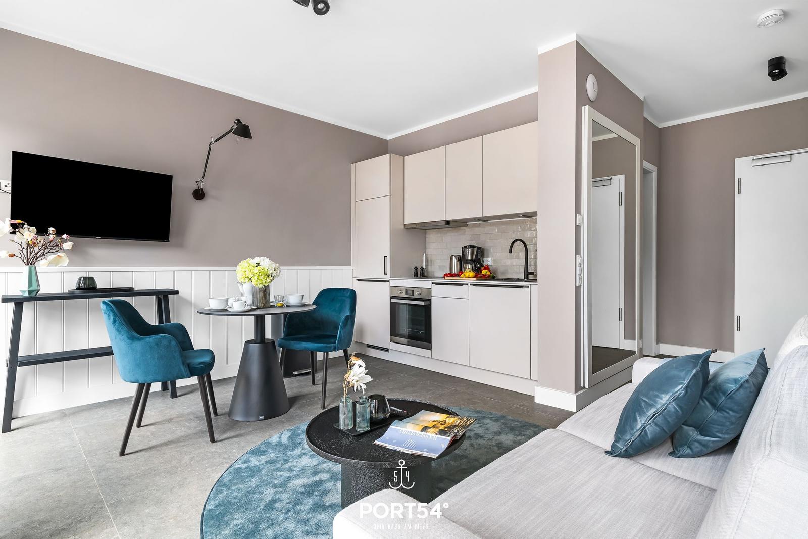 Wohnbereich mit Küche, Sofa und Esstisch. Blaue Stühle und Fernseher an der Wand.