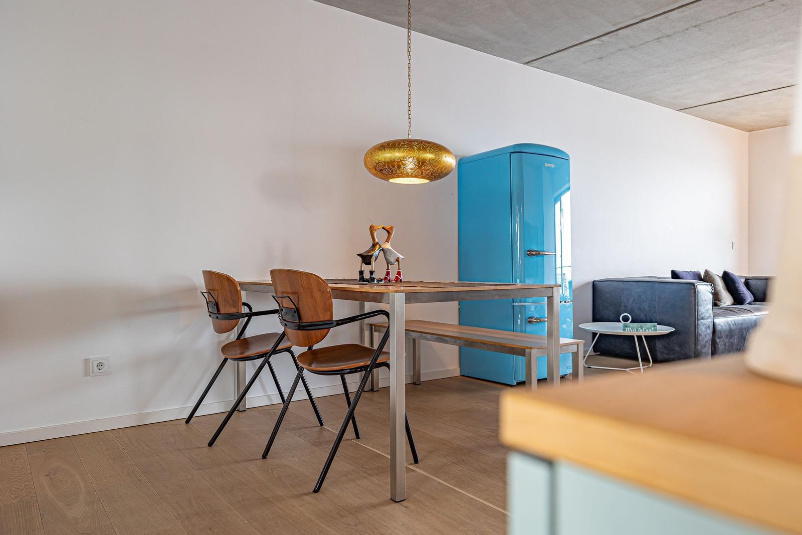 Essbereich mit Tisch, Stühlen und blauem Kühlschrank. Sofa und goldener Hängelampe sind sichtbar.