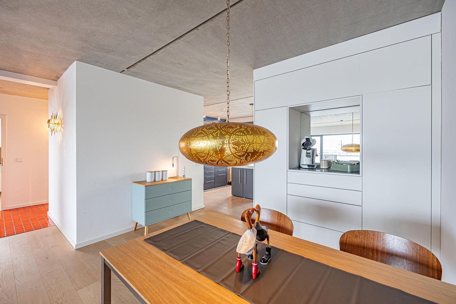 Essbereich mit Holztisch, Stühlen und goldener Hängelampe. Weiße Schrankwand und Küchenbereich im Hintergrund.