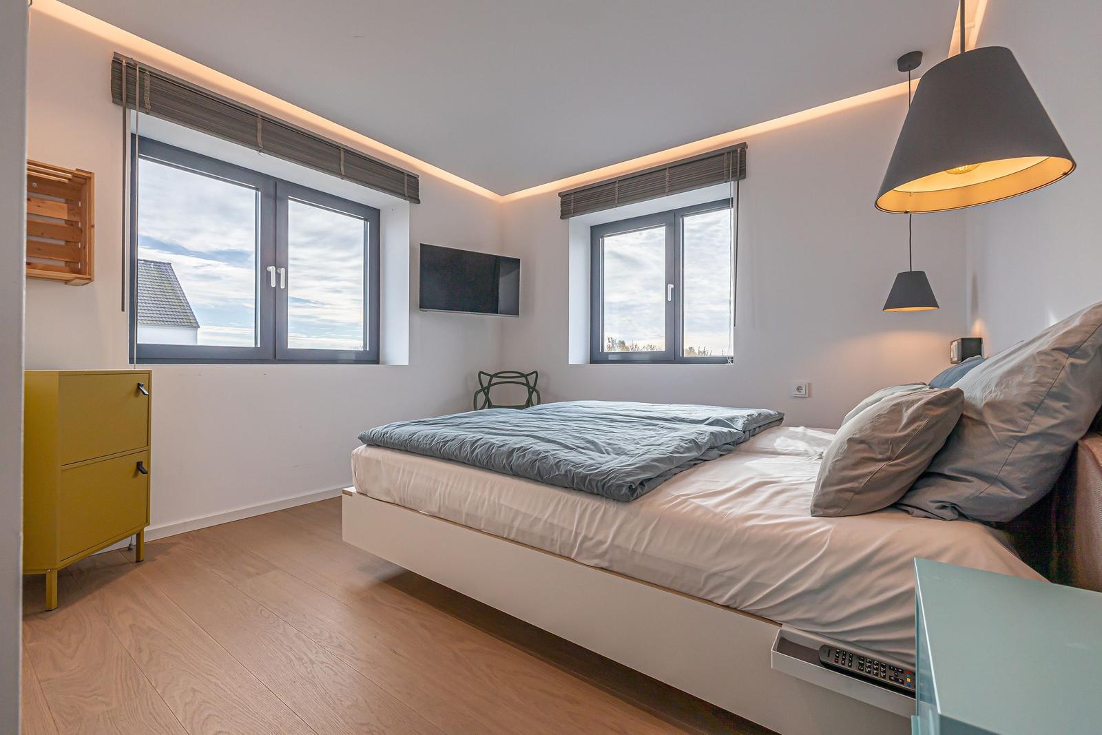 Schlafzimmer mit Bett, Fenstern und TV. Holzboden und gelber Schrank.