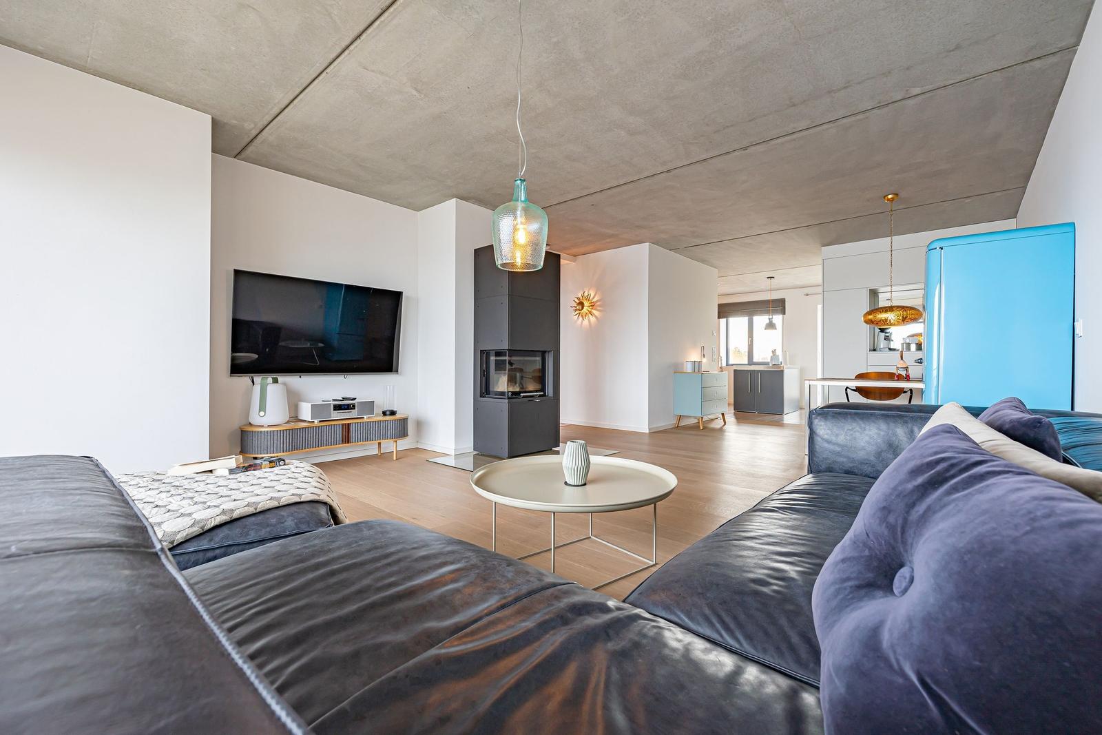 Wohnzimmer mit Sofa, Fernseher und Kamin. Küche mit blauem Kühlschrank sichtbar.