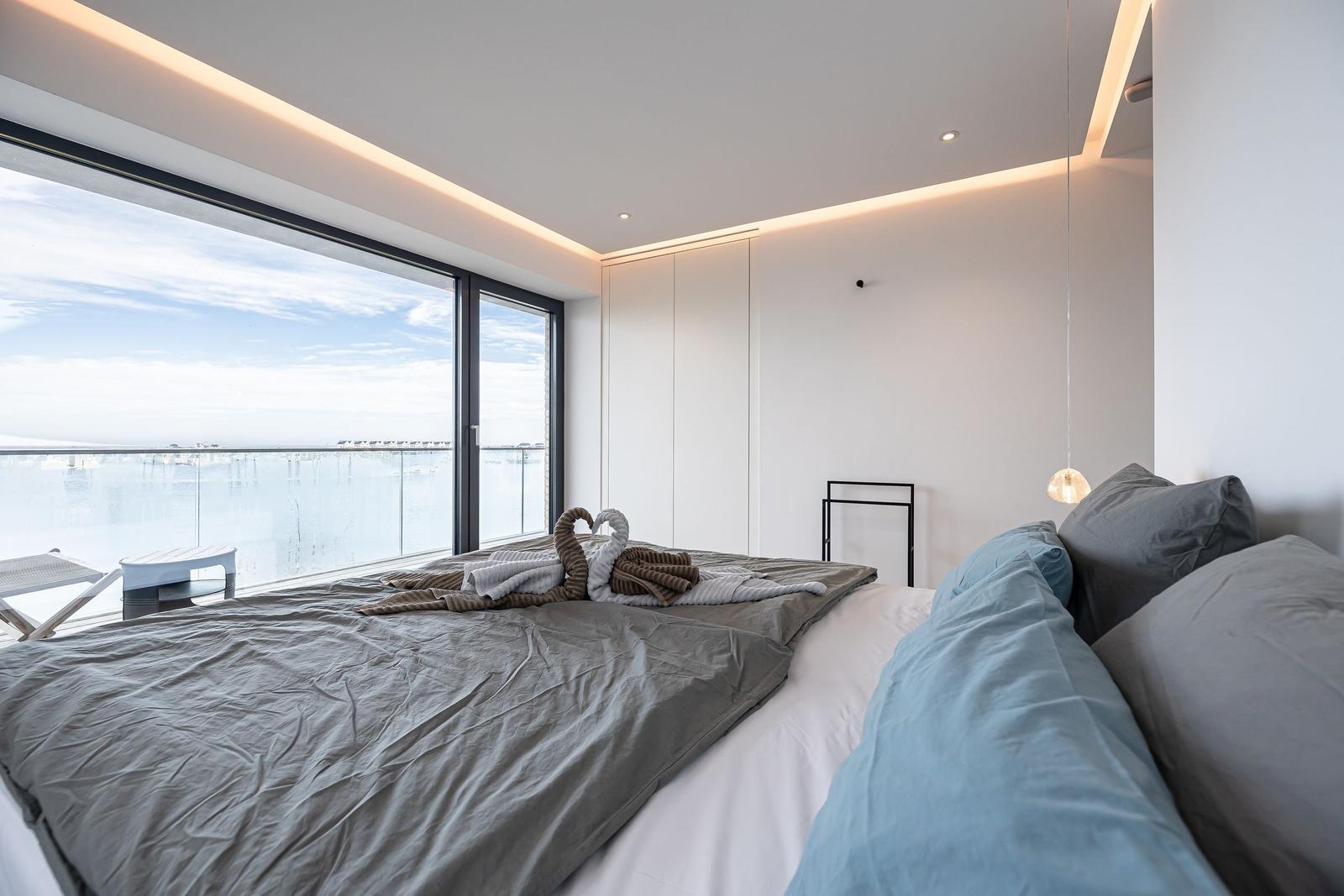 Schlafzimmer mit großem Fenster und Meerblick. Bett mit Kissen und Handtuchschwanen.