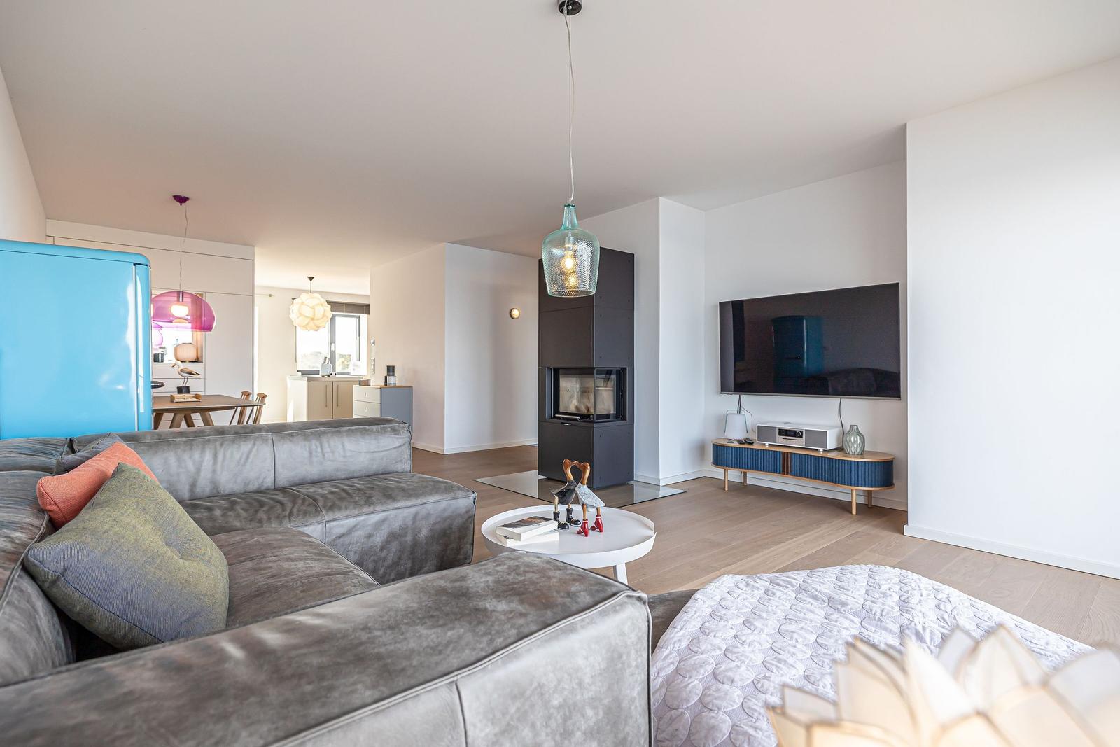 Wohnbereich mit Sofa, Fernseher und Kaminofen. Blauer Kühlschrank und Holzboden sichtbar.