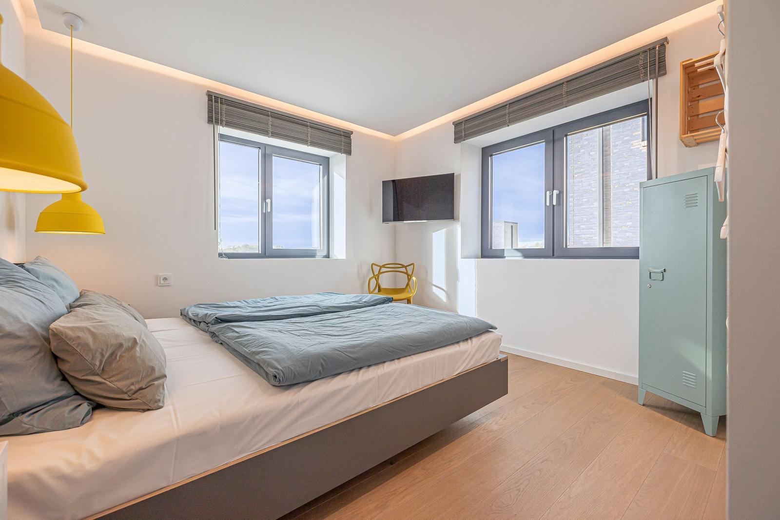 Schlafzimmer mit Bett, Fenstern, Kleiderschrank und Deckenleuchte.