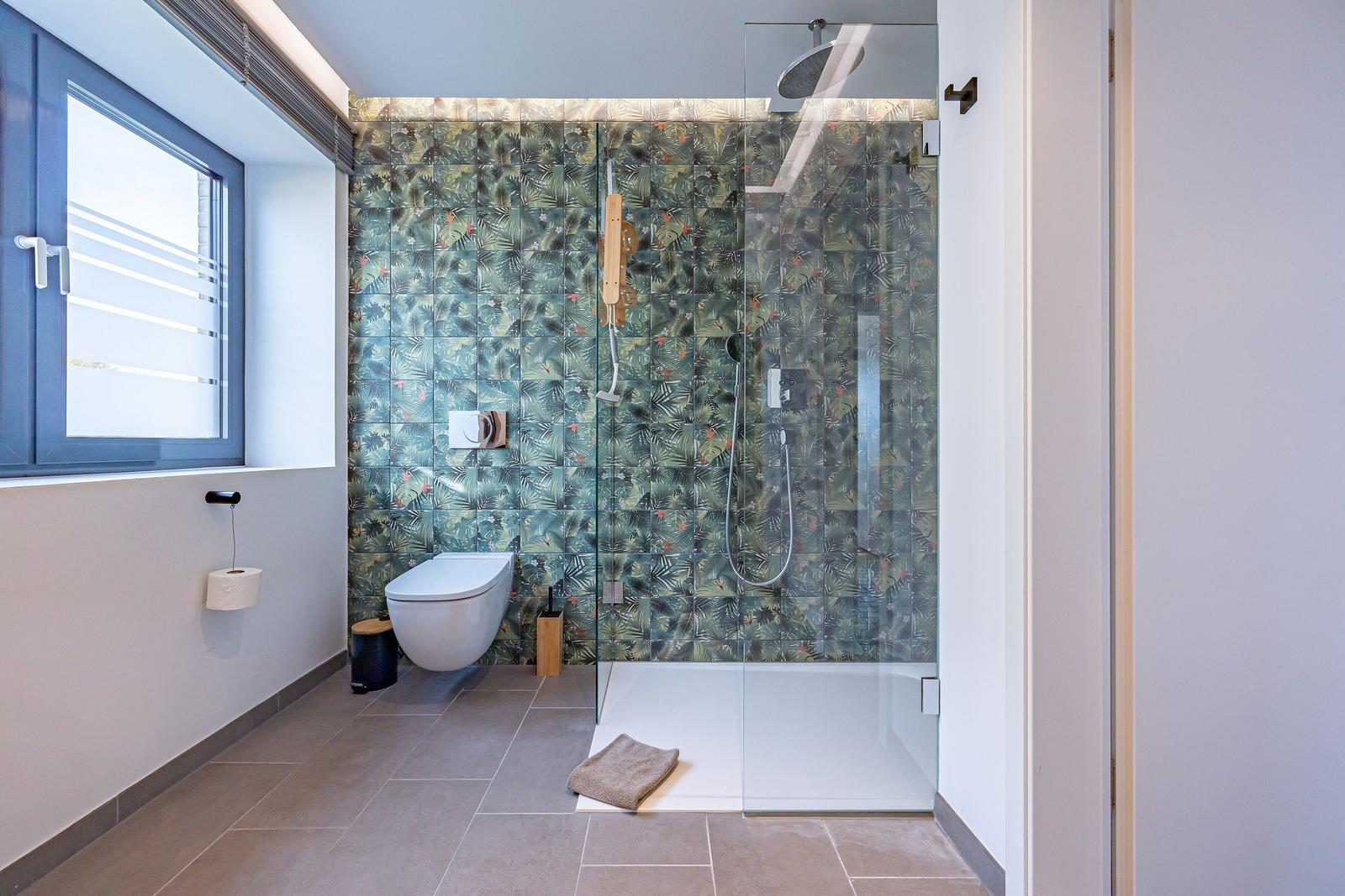Badezimmer mit Dusche, WC und Fenster. Wandfliesen mit tropischem Motiv.
