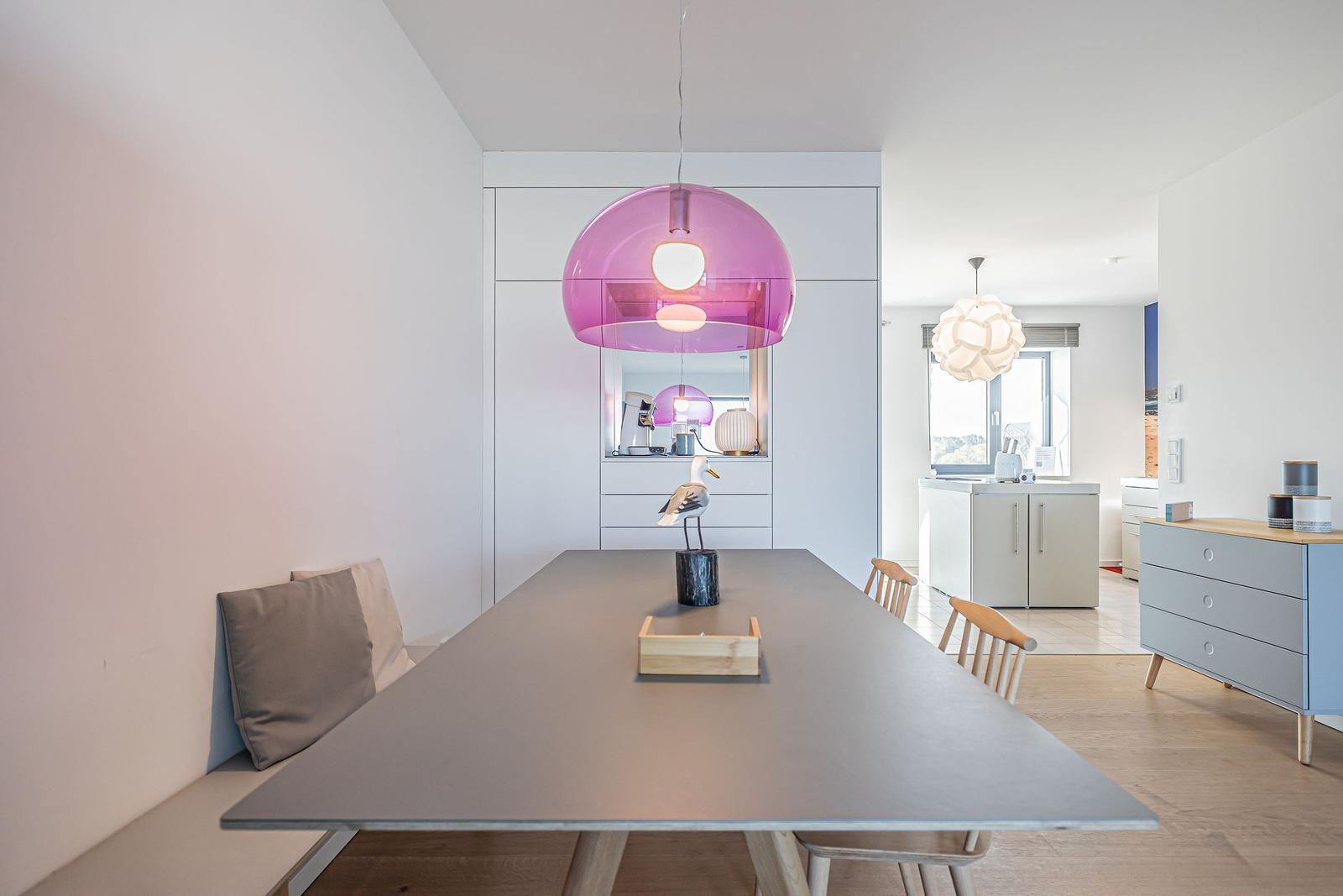 Esszimmer mit langer Tischplatte, pinker Hängelampe und Sitzbank an der Wand.