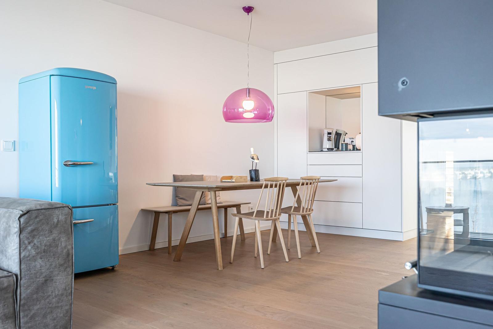 Essbereich mit Holztisch, Stühlen und blauem Kühlschrank unter einer rosa Lampe.