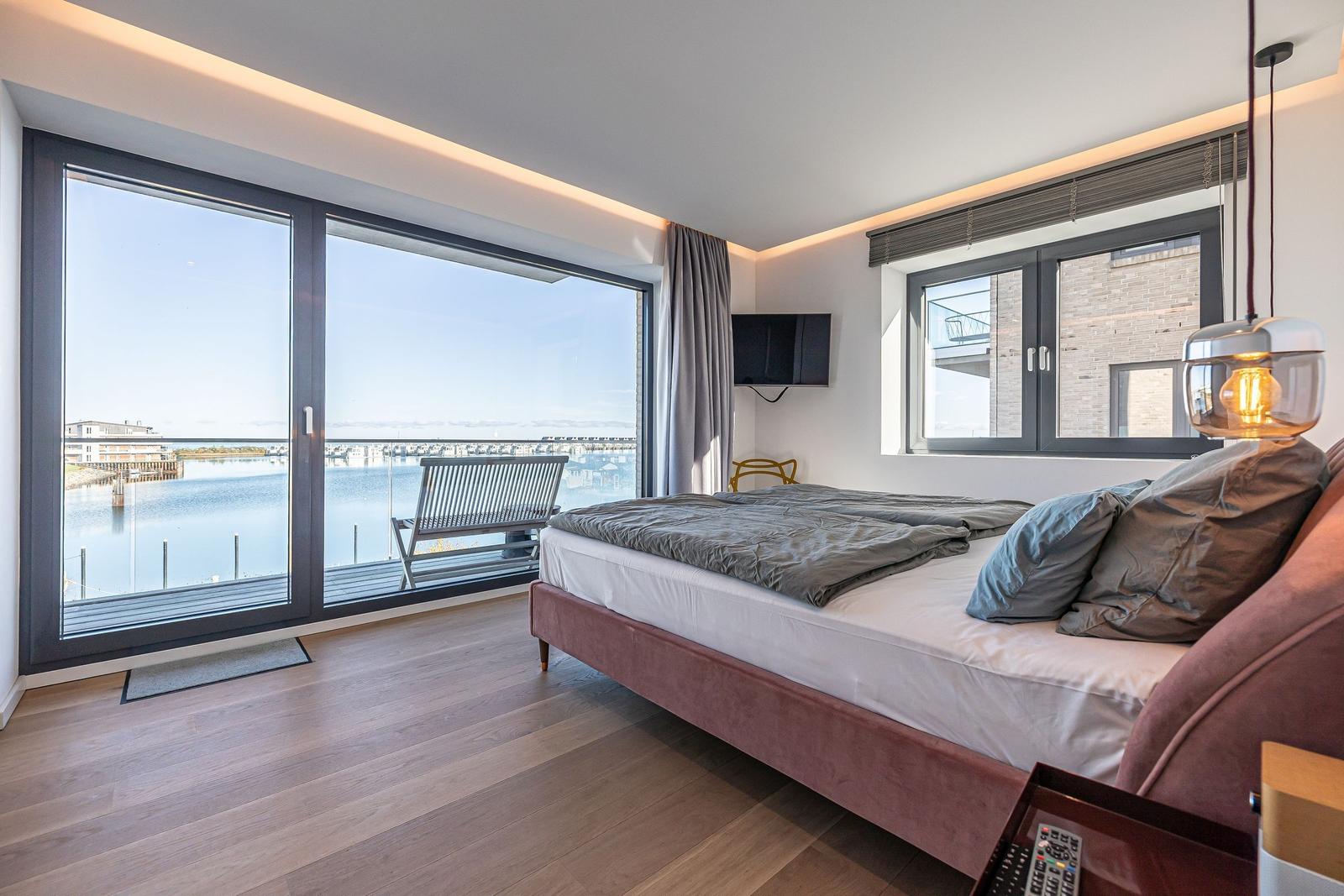 Schlafzimmer mit Blick auf Wasser und Balkon. Bett, TV und moderne Beleuchtung.
