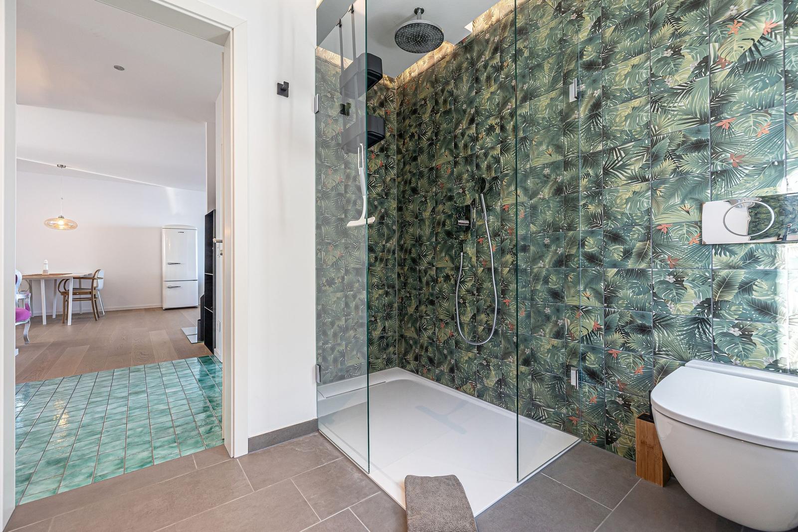 Bad mit Dusche, Toilette und grünem Fliesenboden. Küche und Essbereich sichtbar.