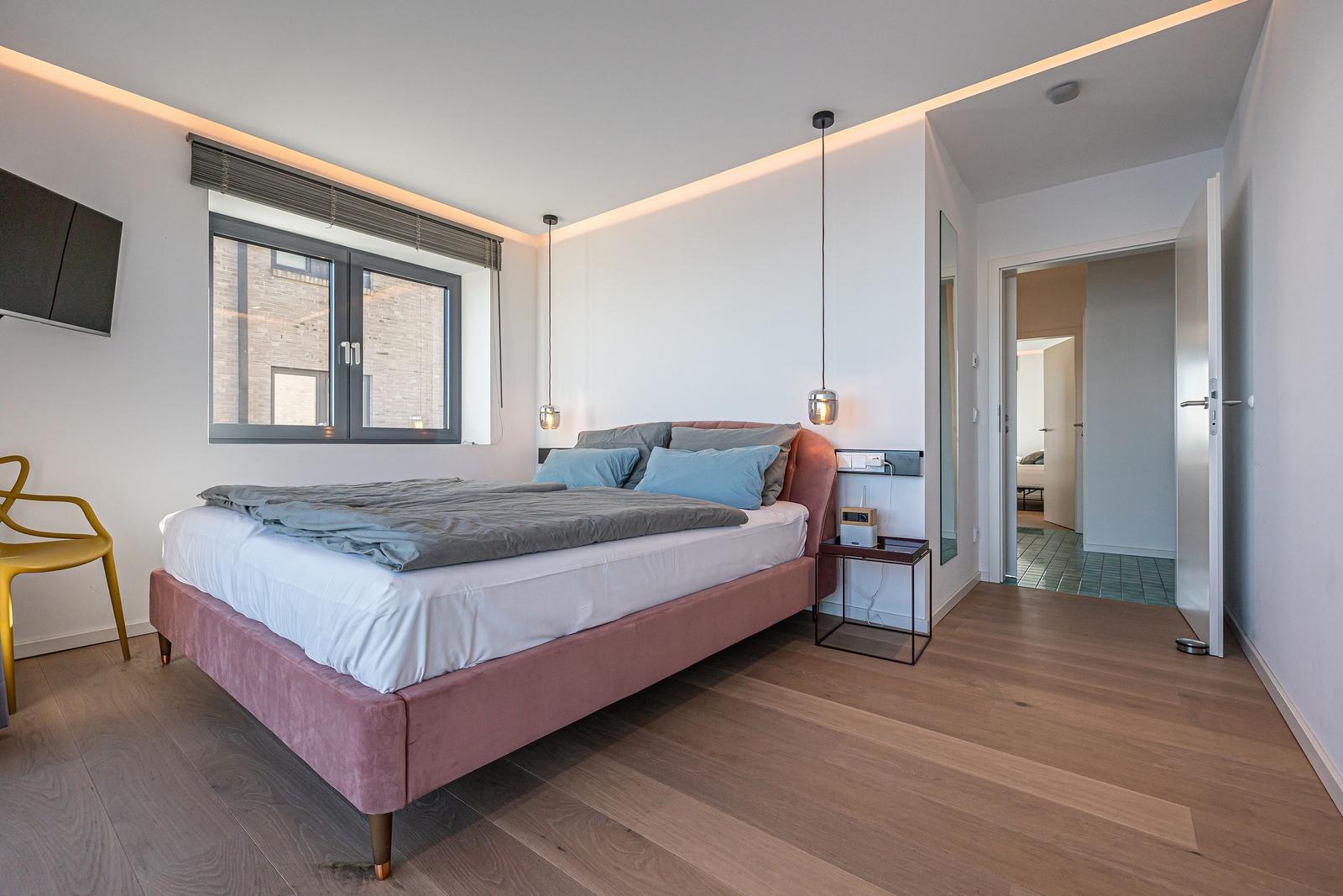 Schlafzimmer mit Bett, Fenster und Holzboden. Gelber Stuhl und Wandspiegel sichtbar.