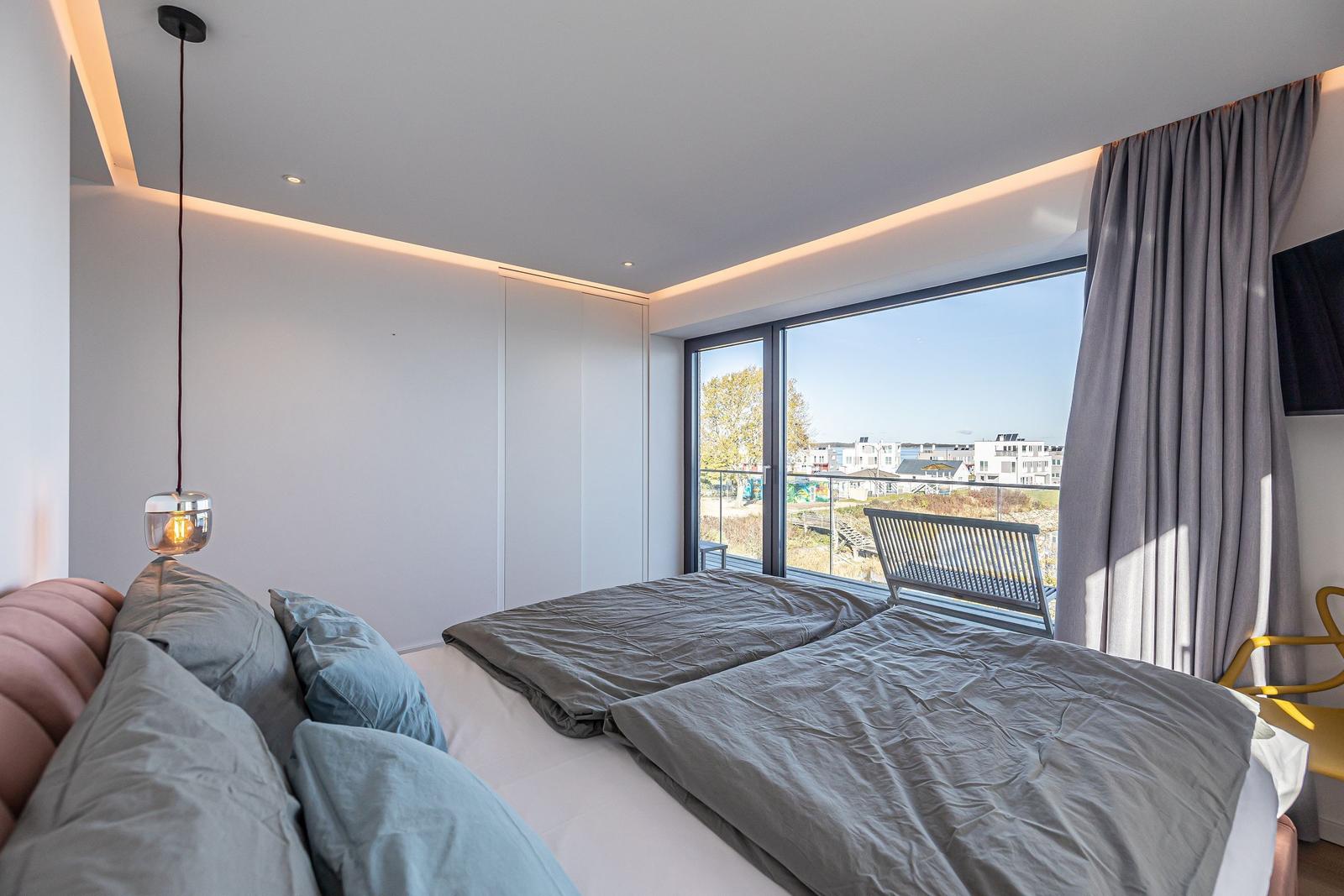 Schlafzimmer mit Blick auf Balkon und Stadt. Bett, Lampe und Vorhänge sind sichtbar.