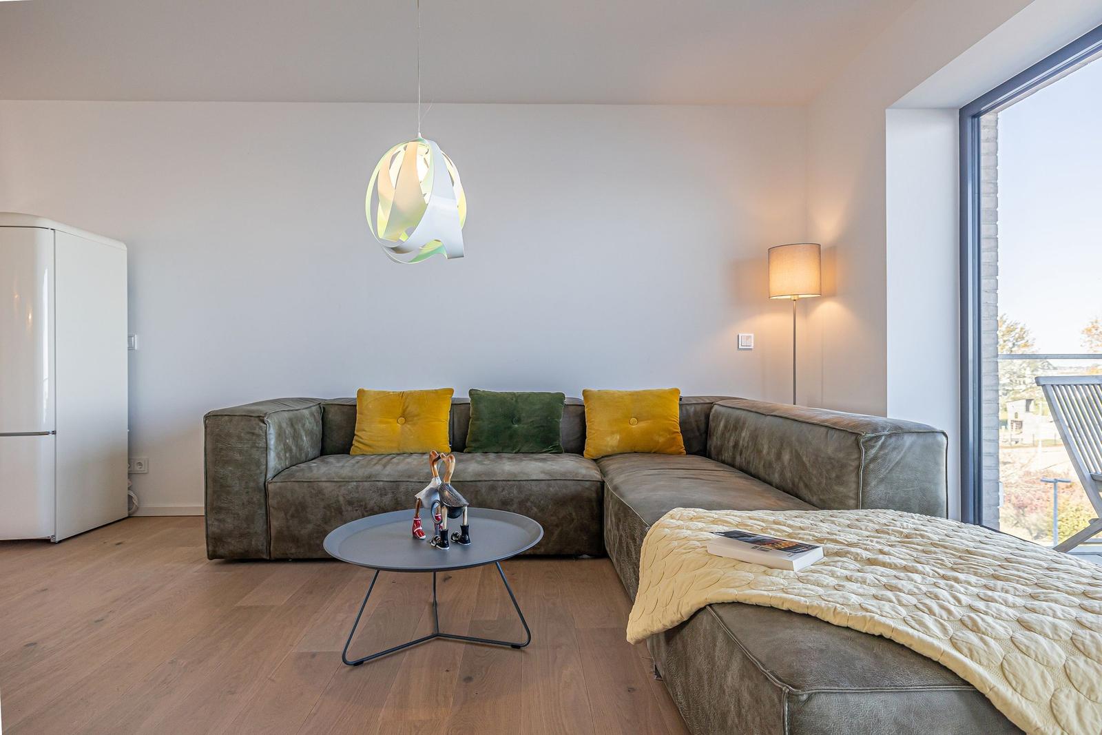 Ein Wohnzimmer mit grauem Sofa, gelben und grünen Kissen, runden Tisch und großen Fenster.