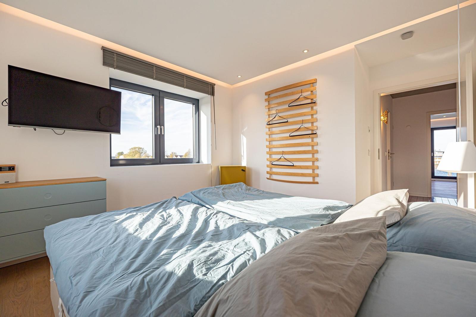 Schlafzimmer mit Bett, TV und Fenster. Holzregal mit Haken an der Wand.
